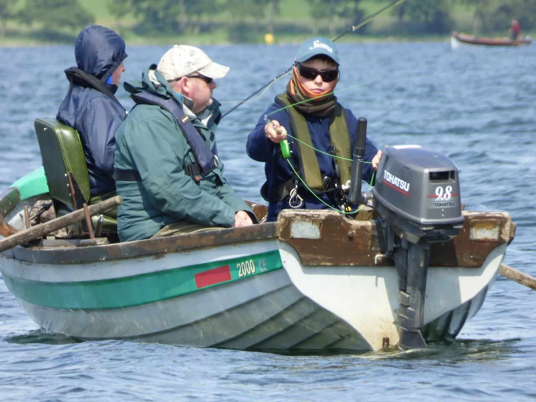 Three men on a small boat fishing on a lake, with another boat visible in the background.
