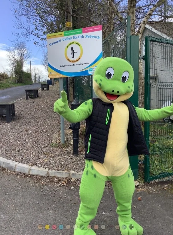 Person in a cartoon frog costume standing outdoors near a fence and holding a sign for Dennett Valley Health Network.
