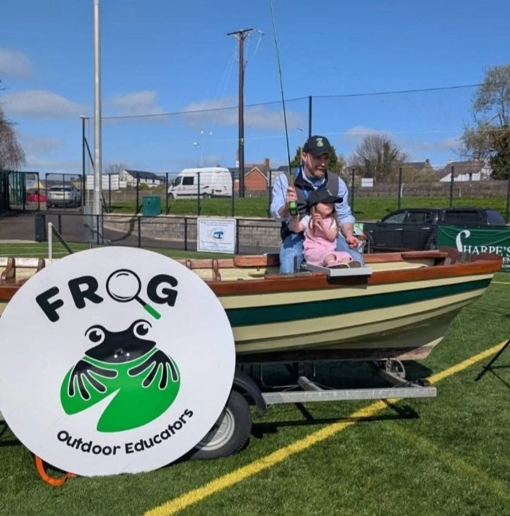 A man and a young girl sitting in a small wooden boat displayed outdoors, with a large round sign reading 'Frog Outdoor Educators' featuring a frog with a magnifying glass. The scene is on a grassy field with a fence, cars, and houses in the background.