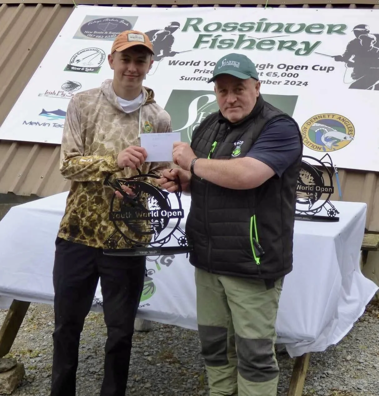 Two men standing in front of a banner at a fishing competition, with one handing over an award to the other, both holding a trophy and an envelope.