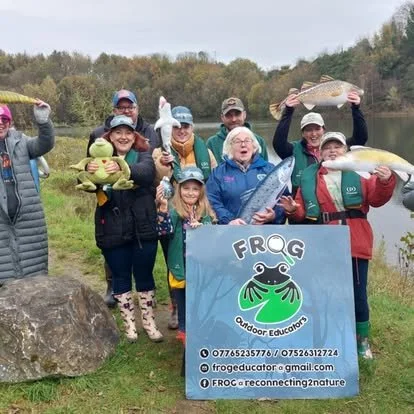 Group of people holding up large fish they caught near a body of water, with a sign that reads 'FROG Outdoor Educators' and features a frog logo.