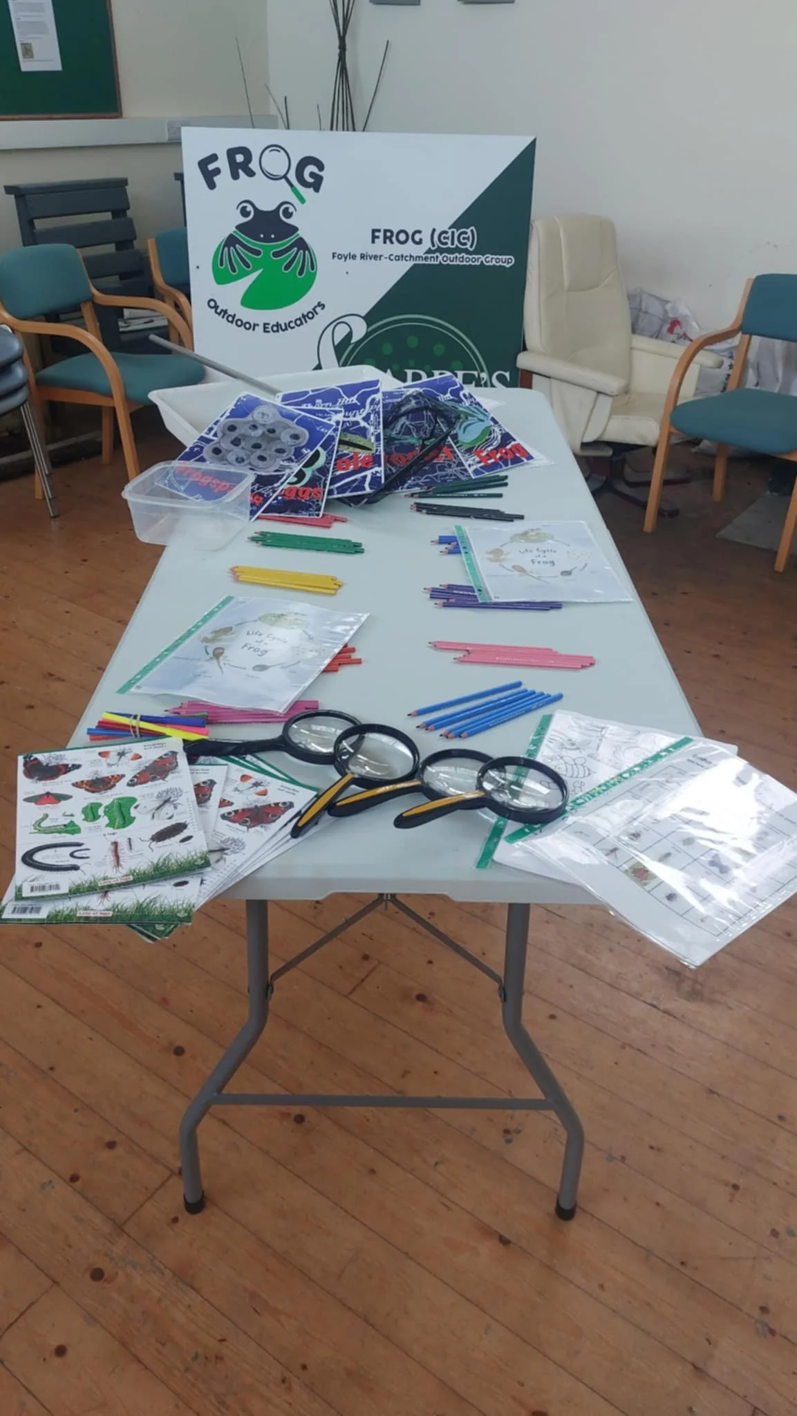 A table with educational materials, coloring pages, plastic magnifying glasses, scissors, plastic straws, and organizing bins, set up for a children’s outdoor educational event with a "FROG" sign and logo in the background.