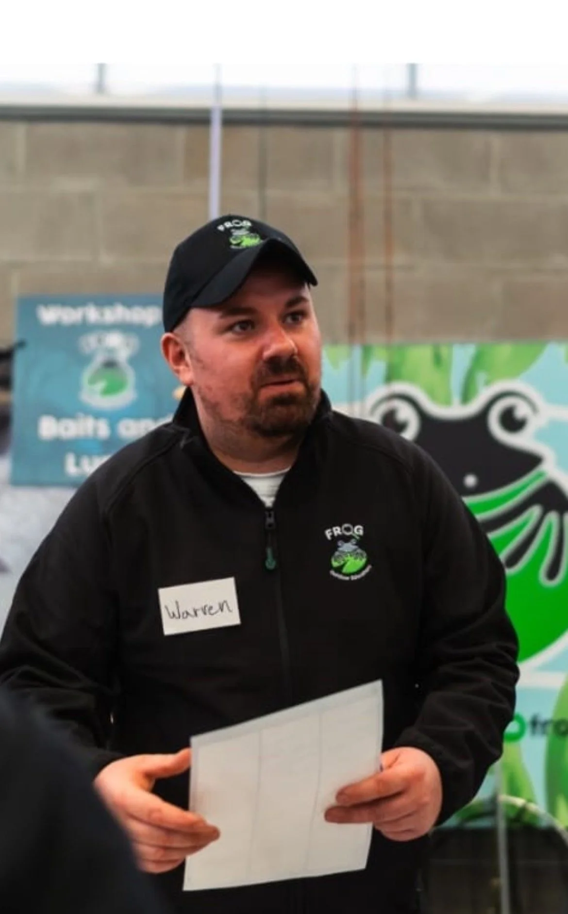 A man with a beard and mustache wearing a black jacket with a frog logo and a black cap, holding a piece of paper, standing in front of a green and black frog logo and a blue banner.