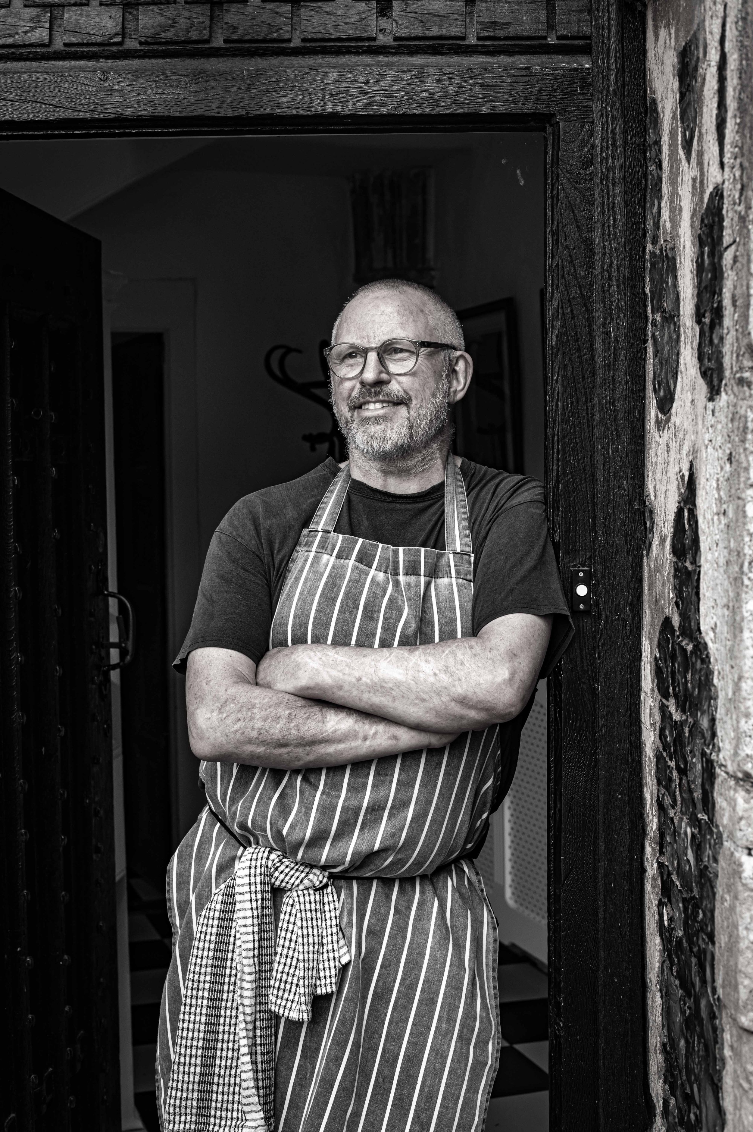 chef peter harrison stands at the door to butley priory, with arms folded wearing a striped apron