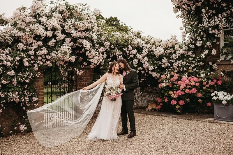 elopement wedding couple standing against a backdrop of pink roses at wedding venue, butley priory, suffolk 