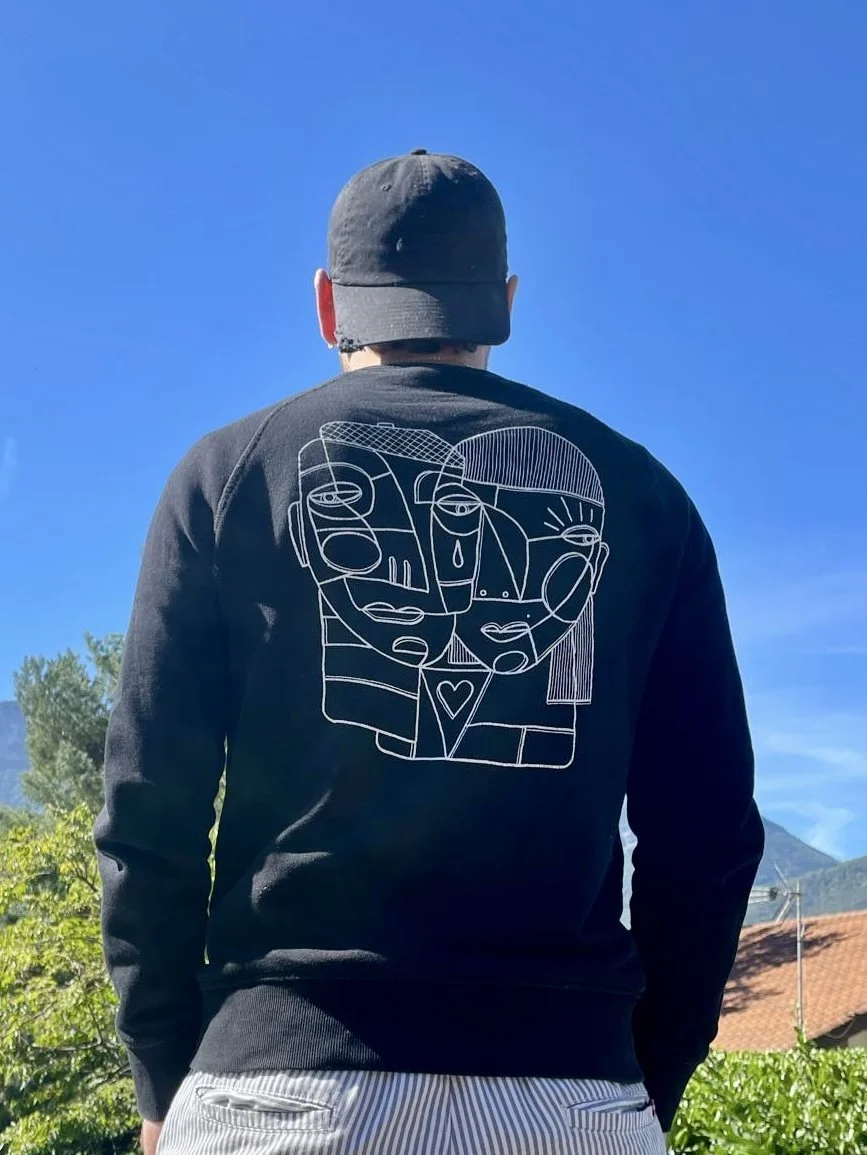 Personne vue de dos portant un sweatshirt noir avec un dessin abstrait de deux visages et un cœur. Personne portant une casquette noire, en extérieur avec un ciel bleu et des arbres en arrière-plan.