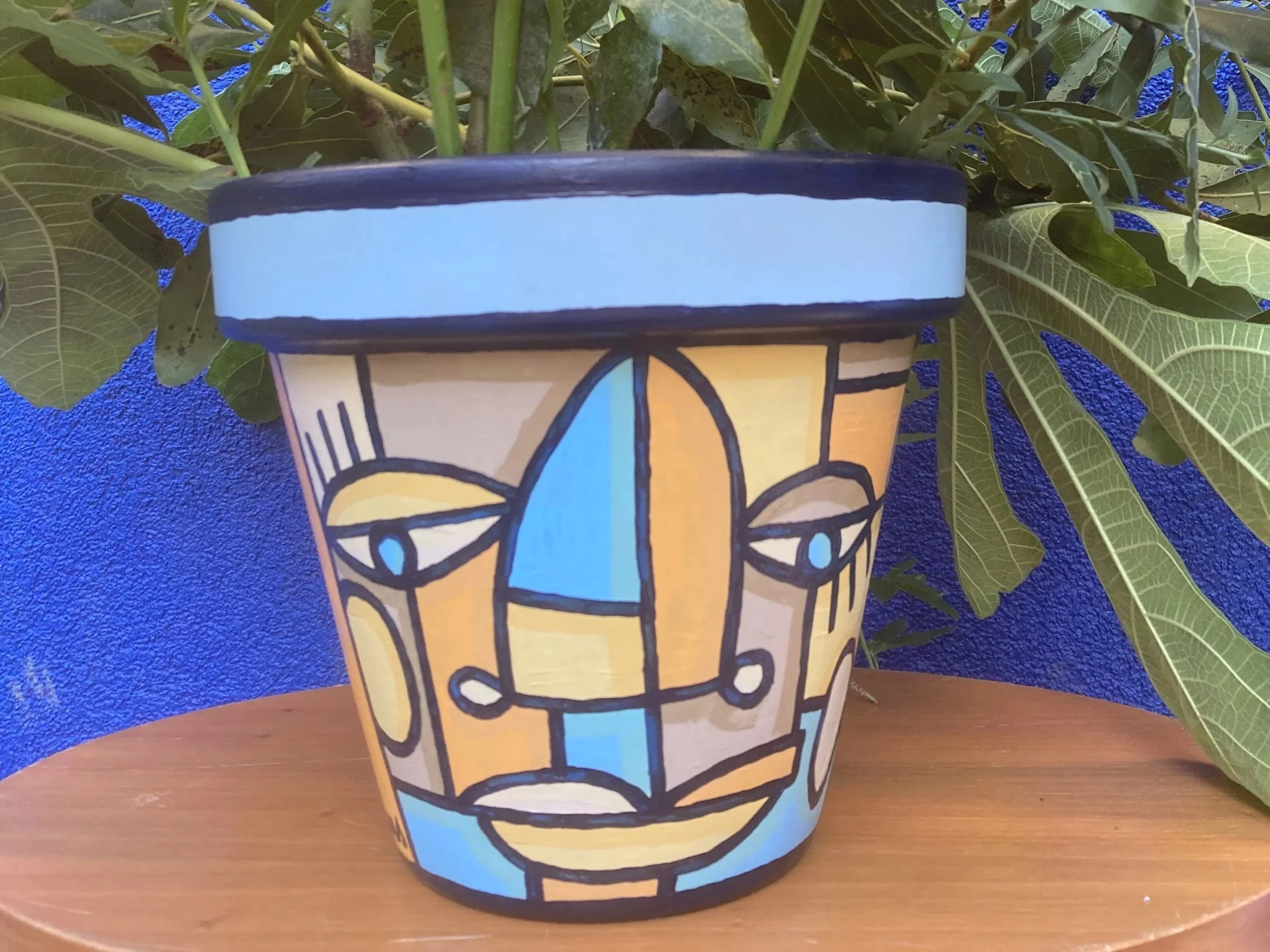 Un pot de fleurs décoré avec une peinture de style abstrait représentant un visage. Le pot est posé sur une table en bois avec des feuilles vertes en arrière-plan et un mur bleu vif derrière.