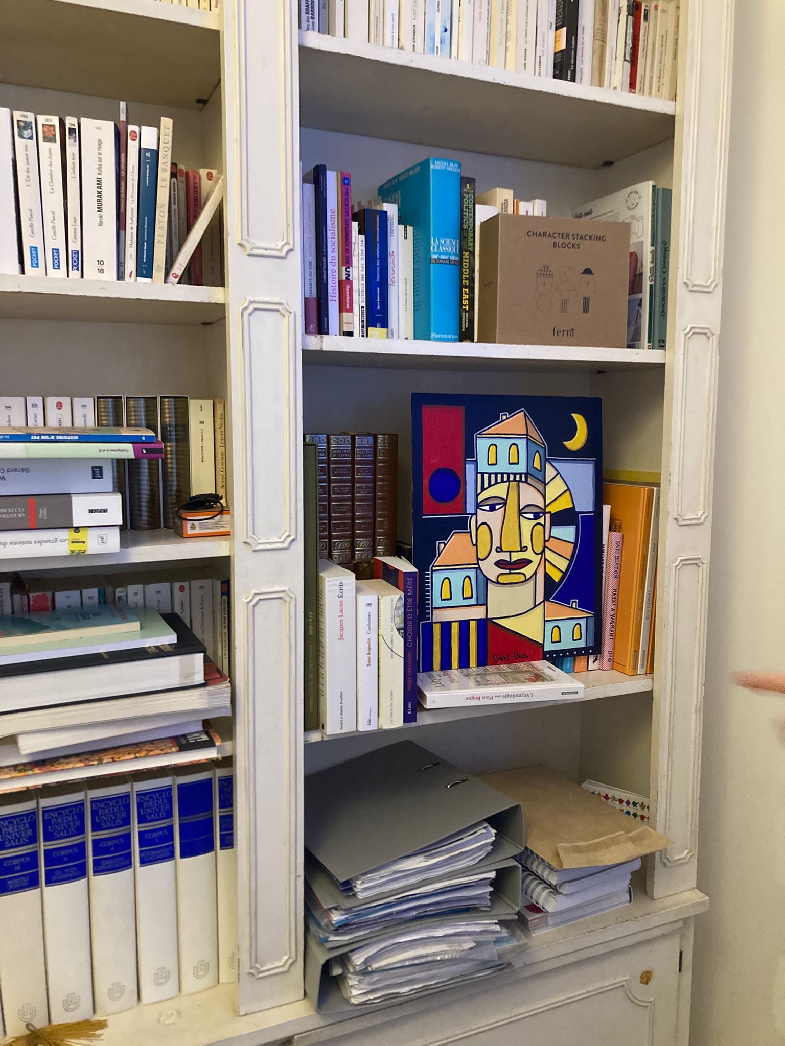 Une bibliothèque avec des livres empilés et rangés, un classeur contenant des papiers empilés, un tableau avec une illustration abstraite de visages et bâtiments, ainsi que quelques boîtes de conserve.