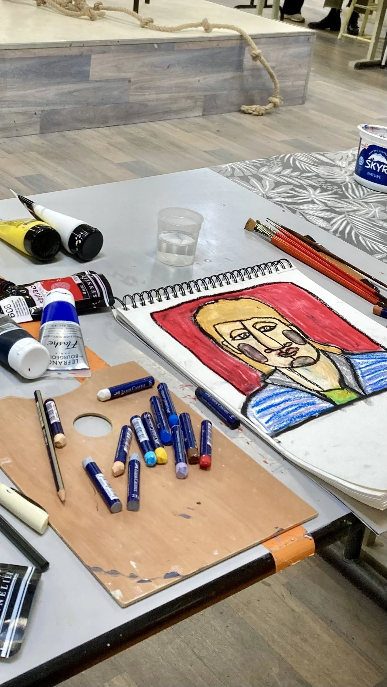 Table d'art avec des tubes de peinture, crayons de couleur, un dessin d'un portrait de femme au style dessin animé, et un tableau derrière avec une corde, dans un espace intérieur.
