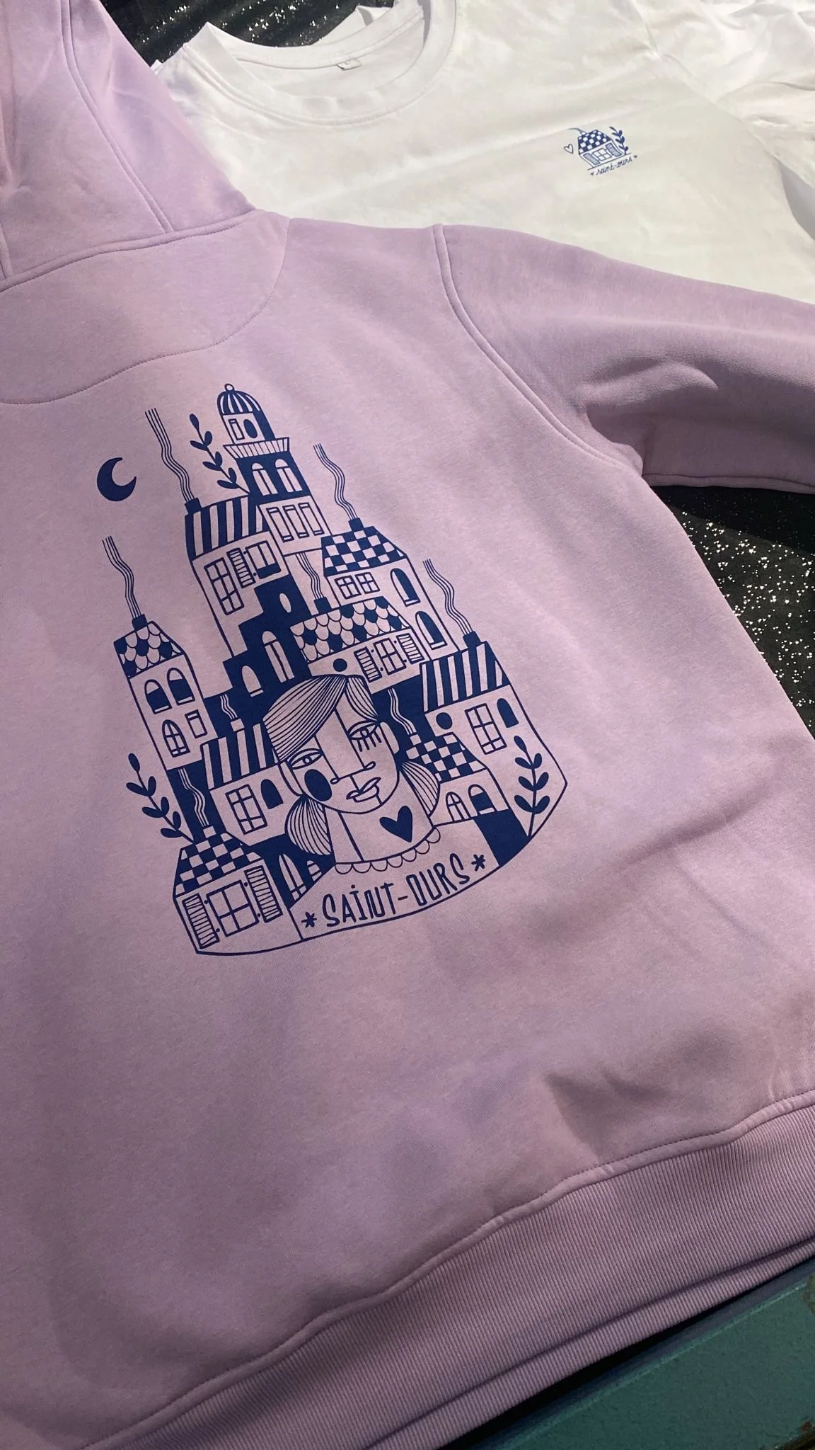 Un sweat-shirt violet clair et un t-shirt blanc disposés à plat, tous deux avec une illustration bleue représentant une ville stylisée avec une femme dans le style d'un dessin minimaliste, et la légende 'Saint-Durs'
