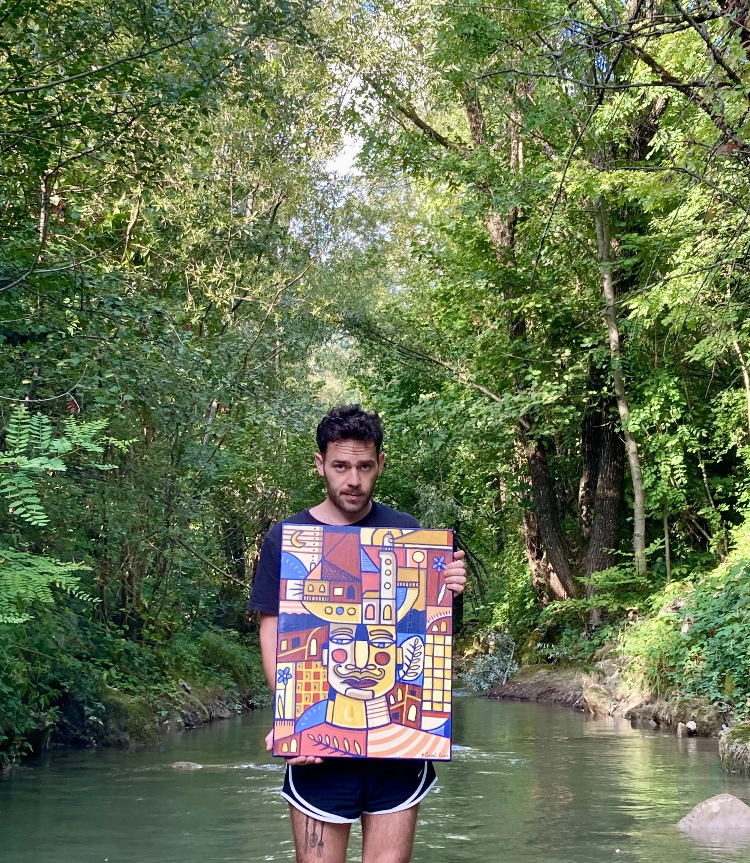 Un homme tient une peinture colorée représentant un visage stylisé avec un paysage urbain en arrière-plan, dans une forêt près d'une rivière.