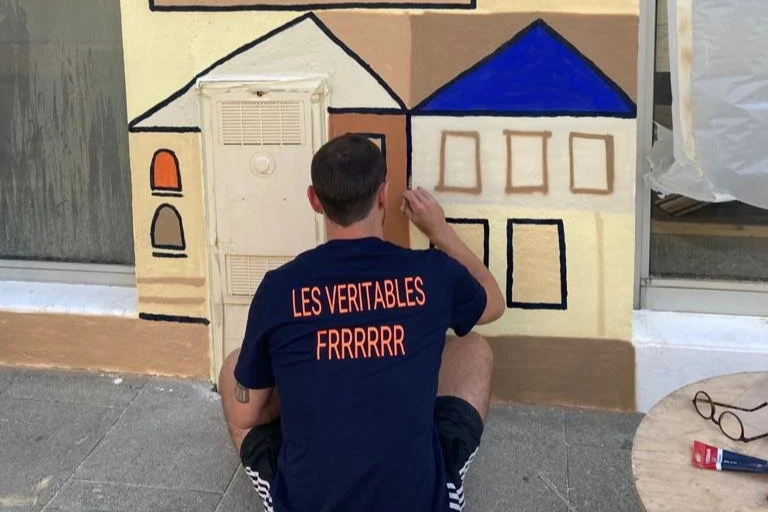 Un homme dessine une maison sur un mur avec de la peinture, en utilisant différentes couleurs et formes.
