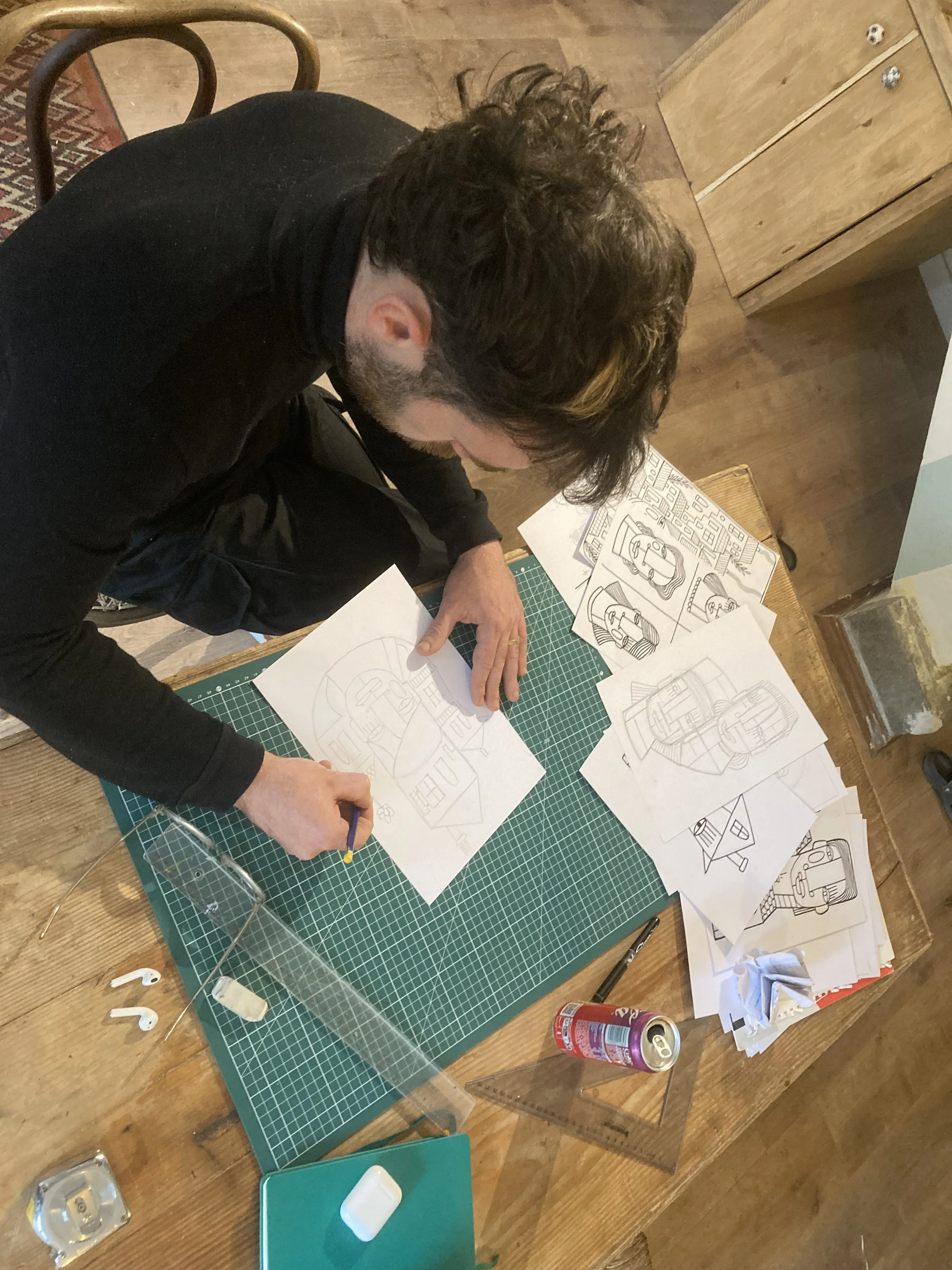 Une personne travaille sur plusieurs dessins à l'écran, avec des outils de dessin, des papiers, des esquisses de voitures et de bâtiments sur une table en bois.