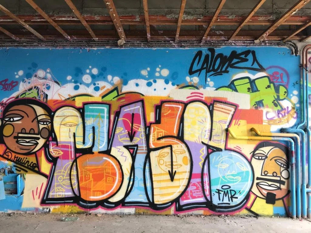 Graffiti coloré avec des personnages stylisés et des lettres stylisées sur un mur, sous un plafond en bois.