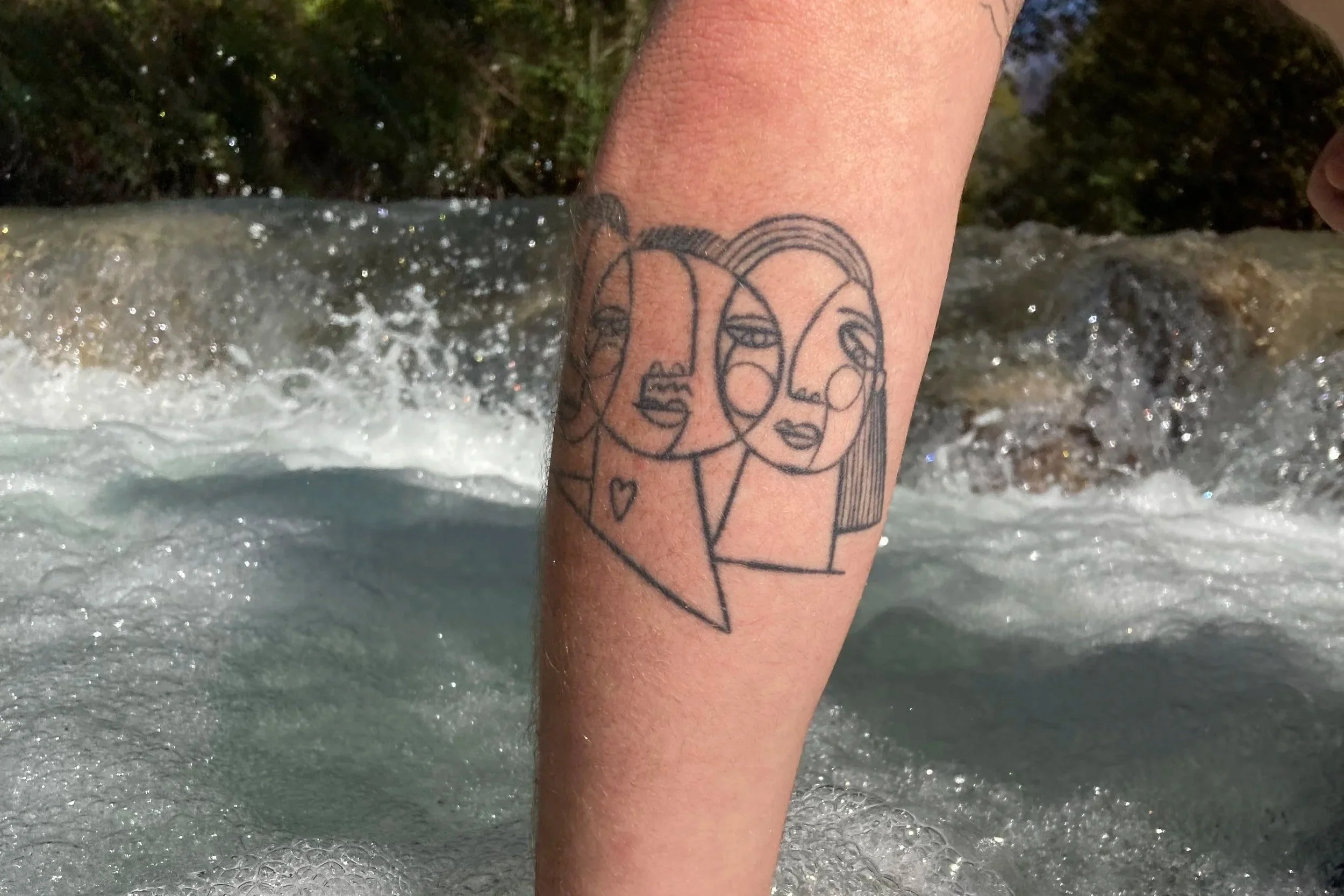 Tatouage minimaliste de deux visages féminins dessinés avec des lignes continues sur une jambe, en arrière-plan une rivière ou une chute d'eau.