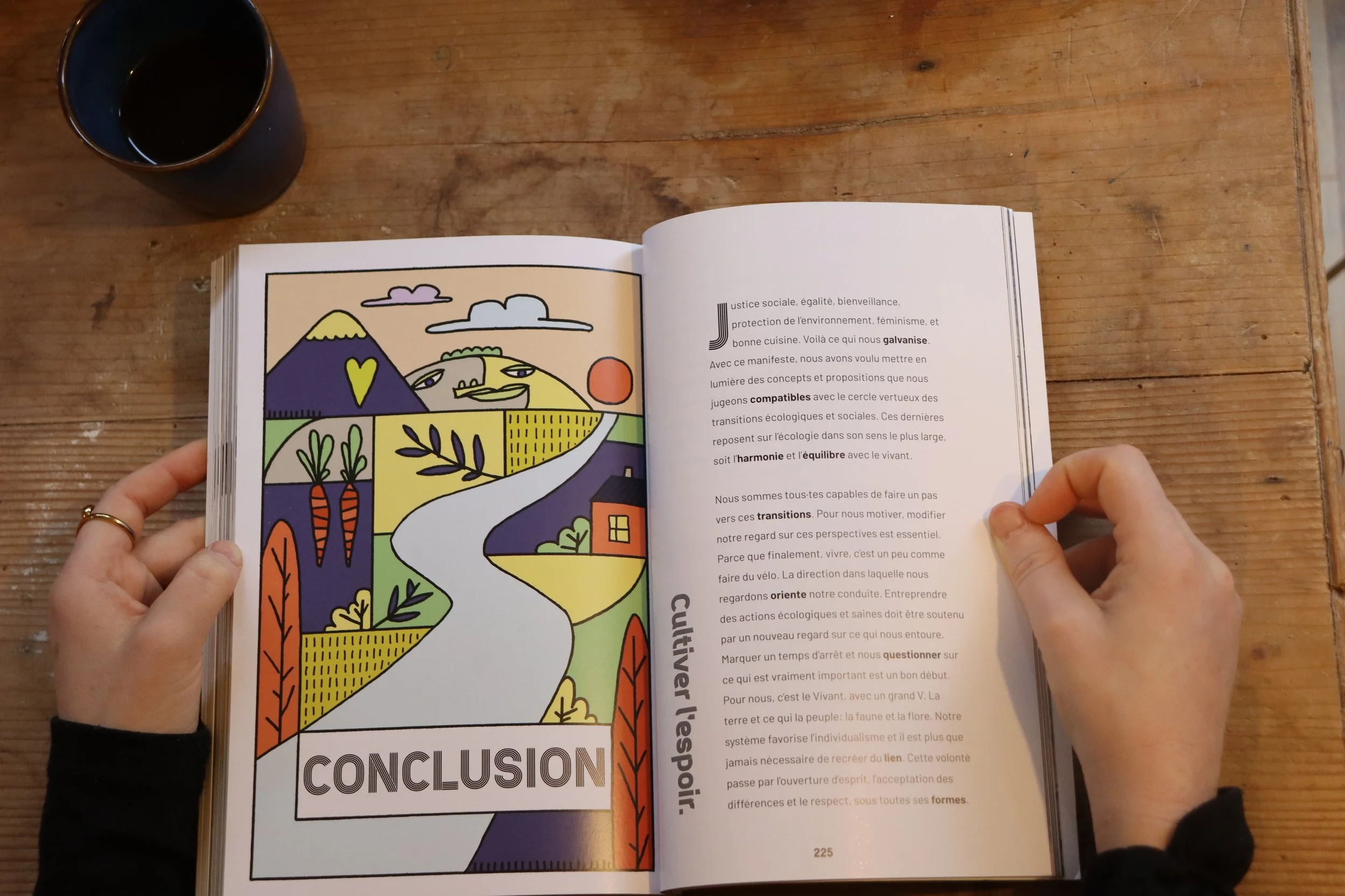 Une personne tient un livre ouvert sur une table en bois, avec la page de gauche illustrée avec une représentation stylisée d'un paysage avec montagnes, arbres, soleil et une route, et la page de droite contenant du texte en français. Une tasse à café noire est visible en haut à gauche.
