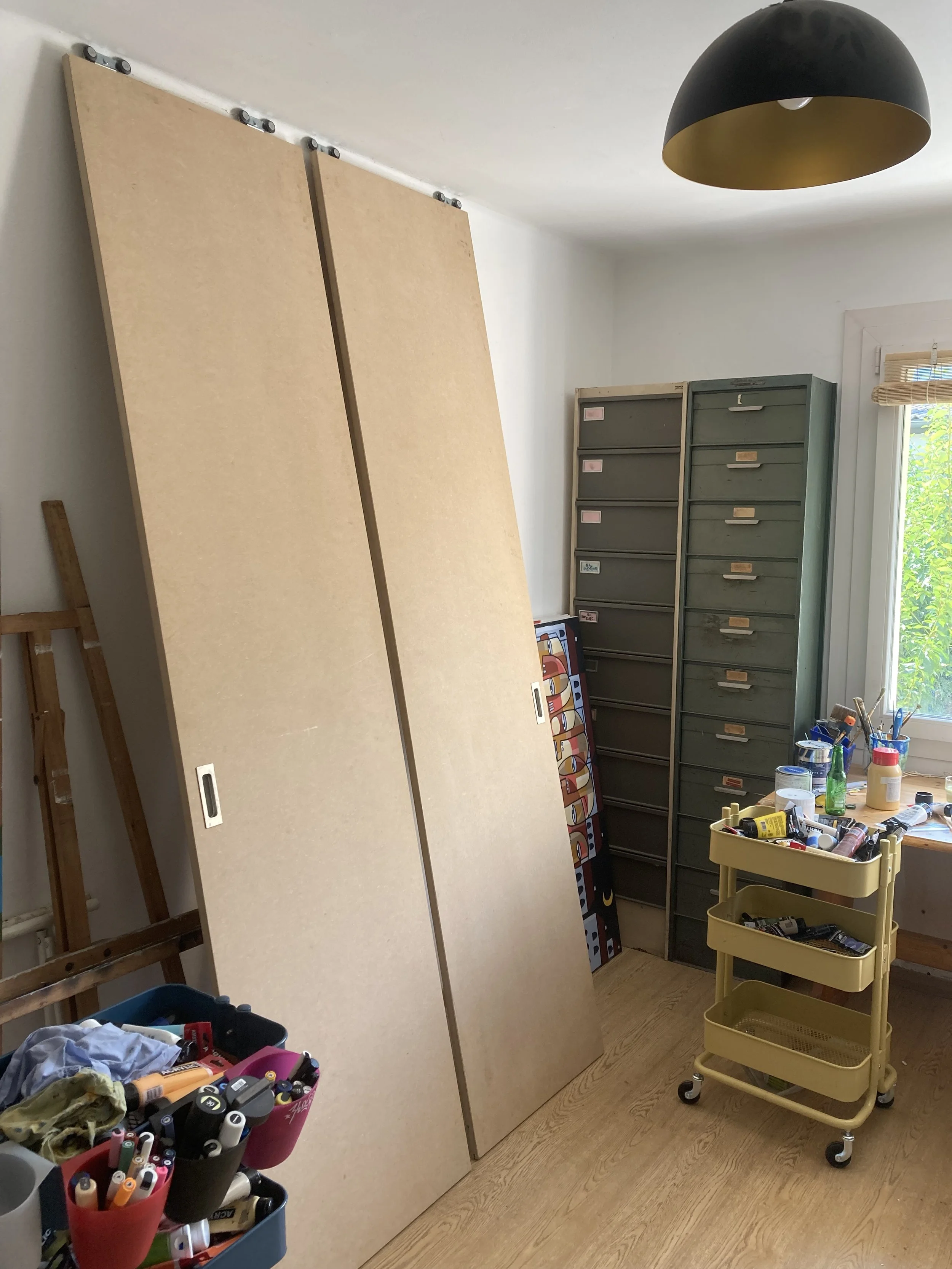 Une pièce de bricolage ou d'art avec deux grands panneaux en bois, une caisse à outils avec des marqueurs, un meuble de rangement métallique, une fenêtre laissant entrer de la lumière.