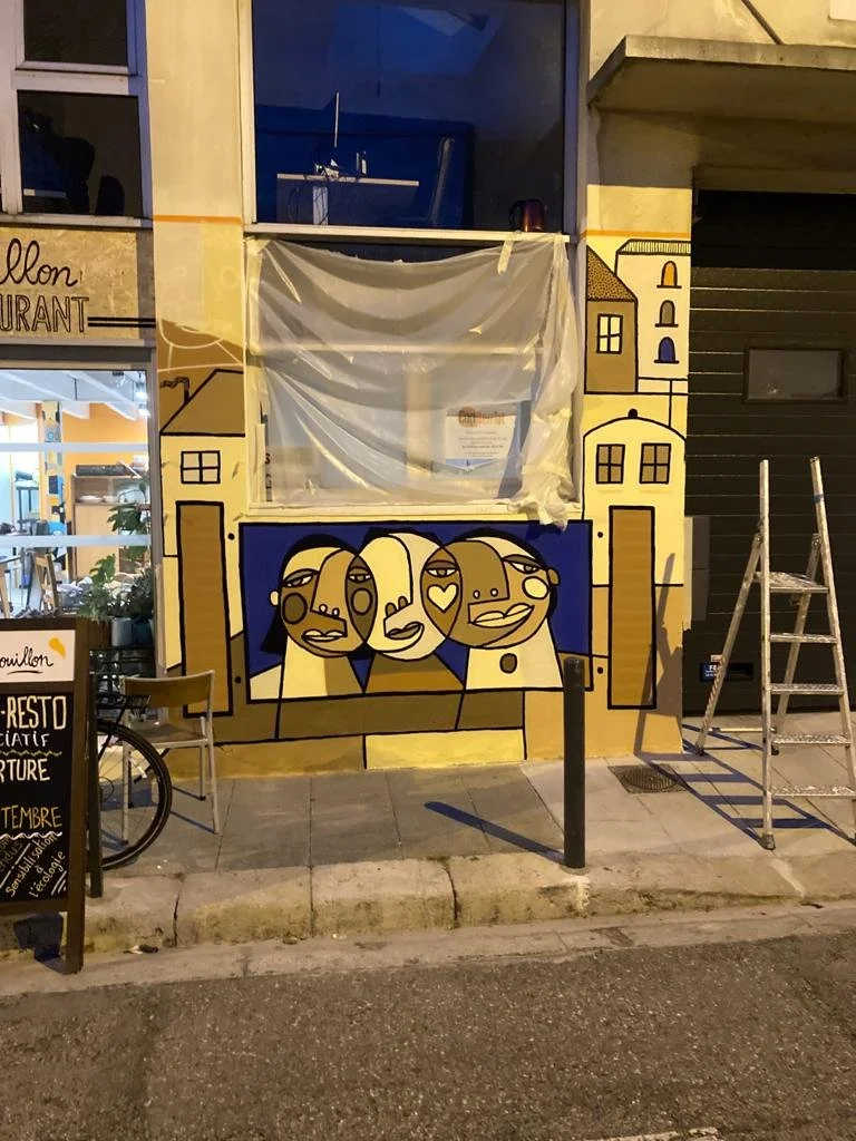 Une fresque murale colorée représentant trois visages stylisés avec des traits abstraits à l'extérieur d'un bâtiment. Il y a aussi une échelle en métal à droite et un tableau d'affichage avec des textes.