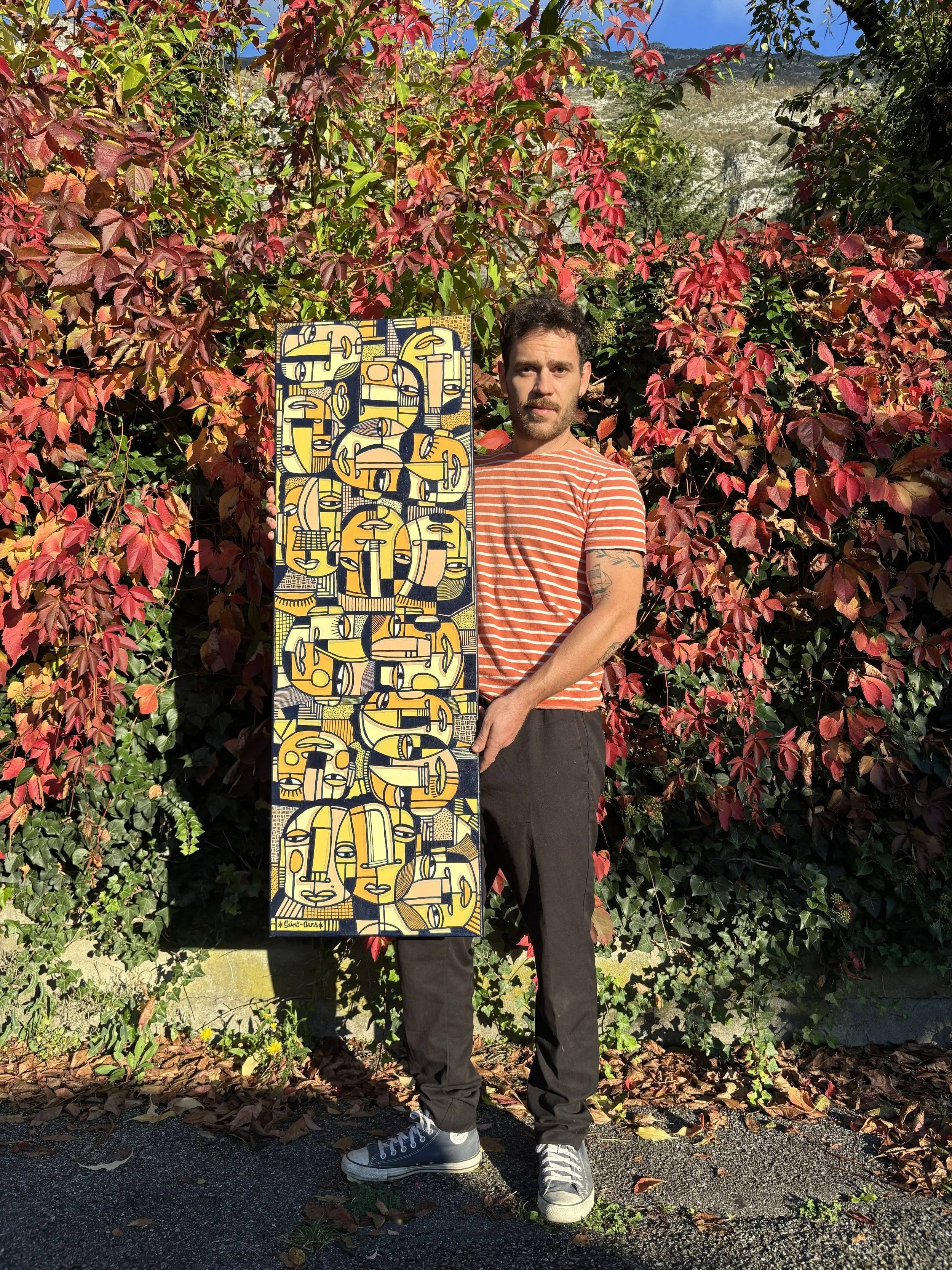 Un homme tient une peinture colorée avec des formes abstraites, se tenant devant un buisson rouge et vert en plein air par temps ensoleillé.