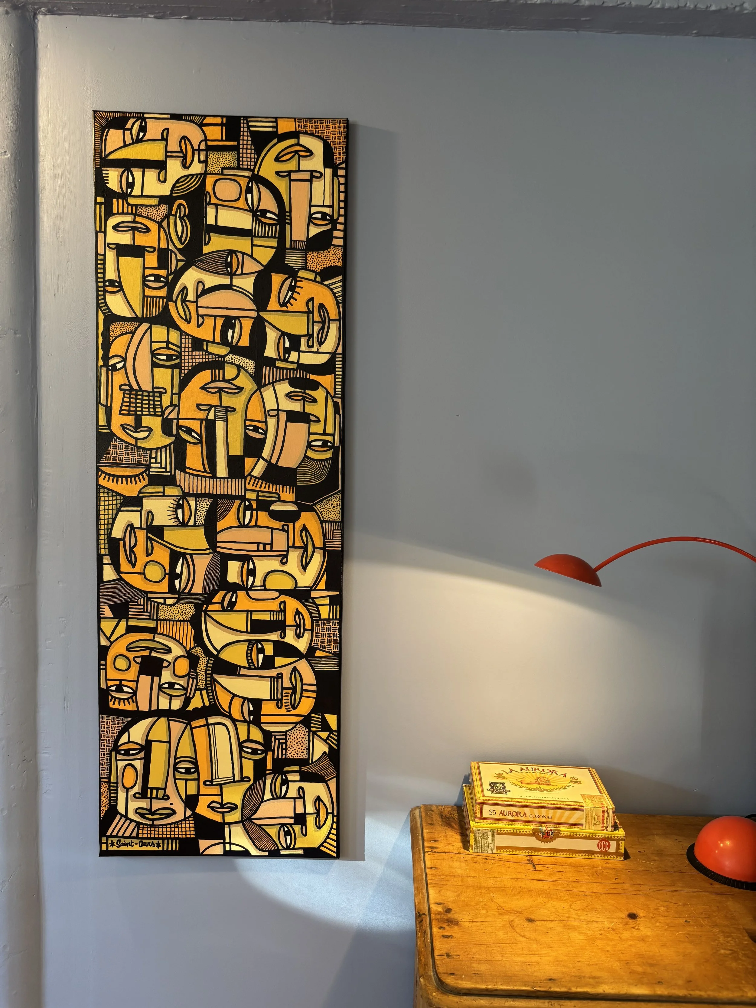 Une peinture abstraite avec des visages stylisés en nuances de jaune, orange, noir et blanc, accrochée à un mur gris.