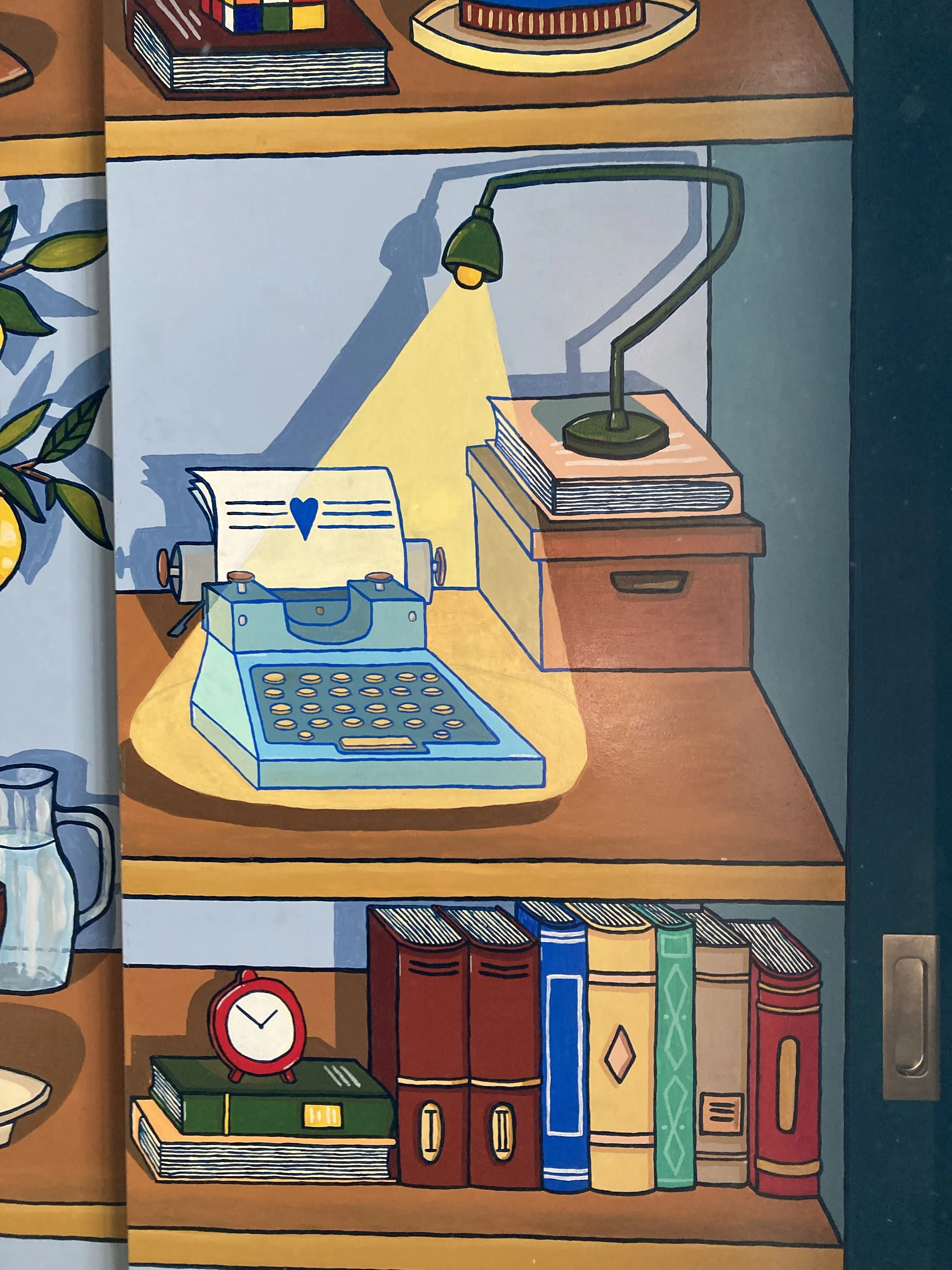 Une étagère avec un bureau, une lampe, des livres, une machine à écrire avec un cœur dessus, une horloge rouge et une cruche à côté.