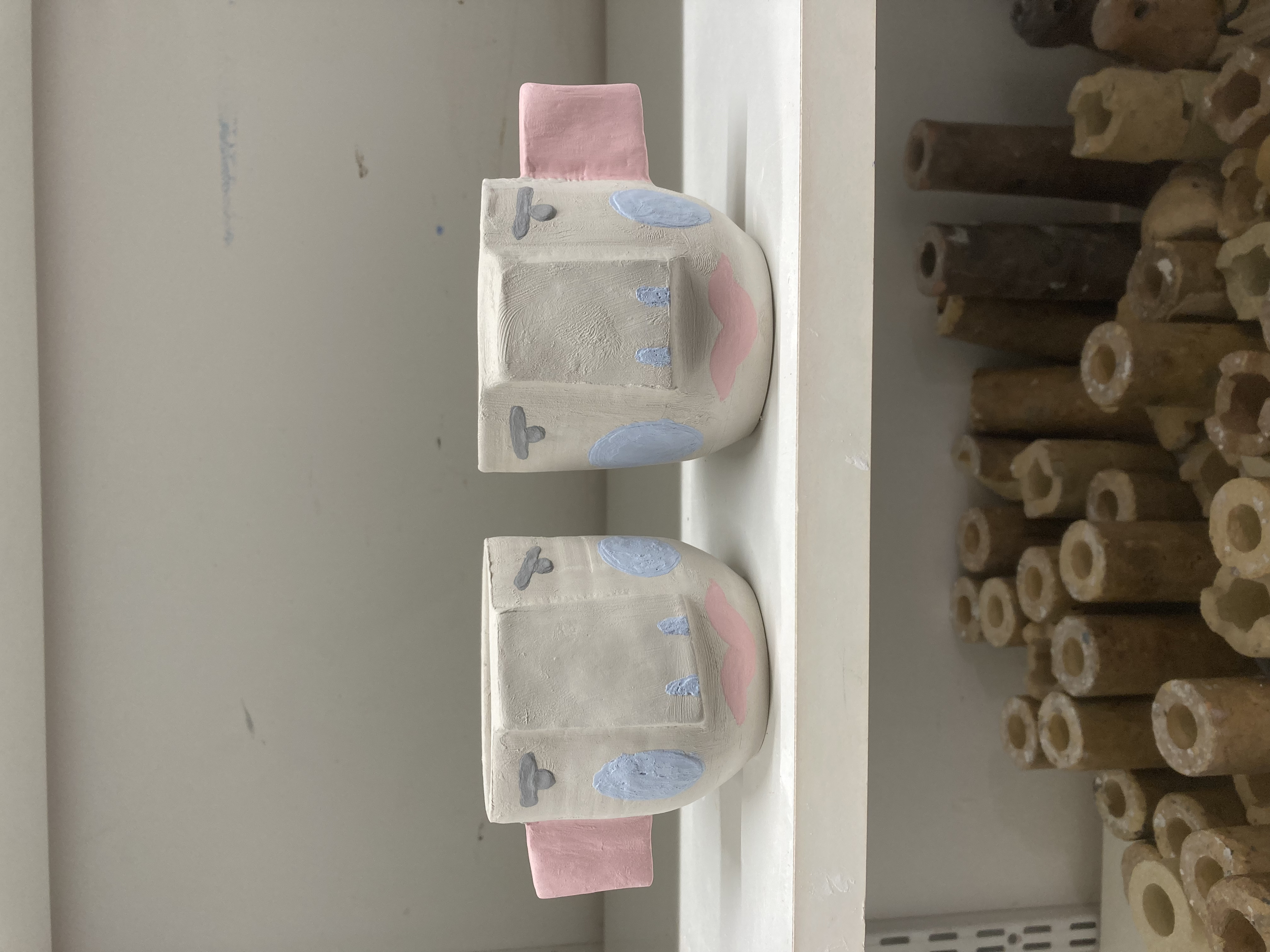 Deux tasses en céramique peintes avec des motifs roses, bleus et gris, placées sur une étagère blanche. À l'arrière-plan, un mur beige, et à droite, un tas de tubes en bois.