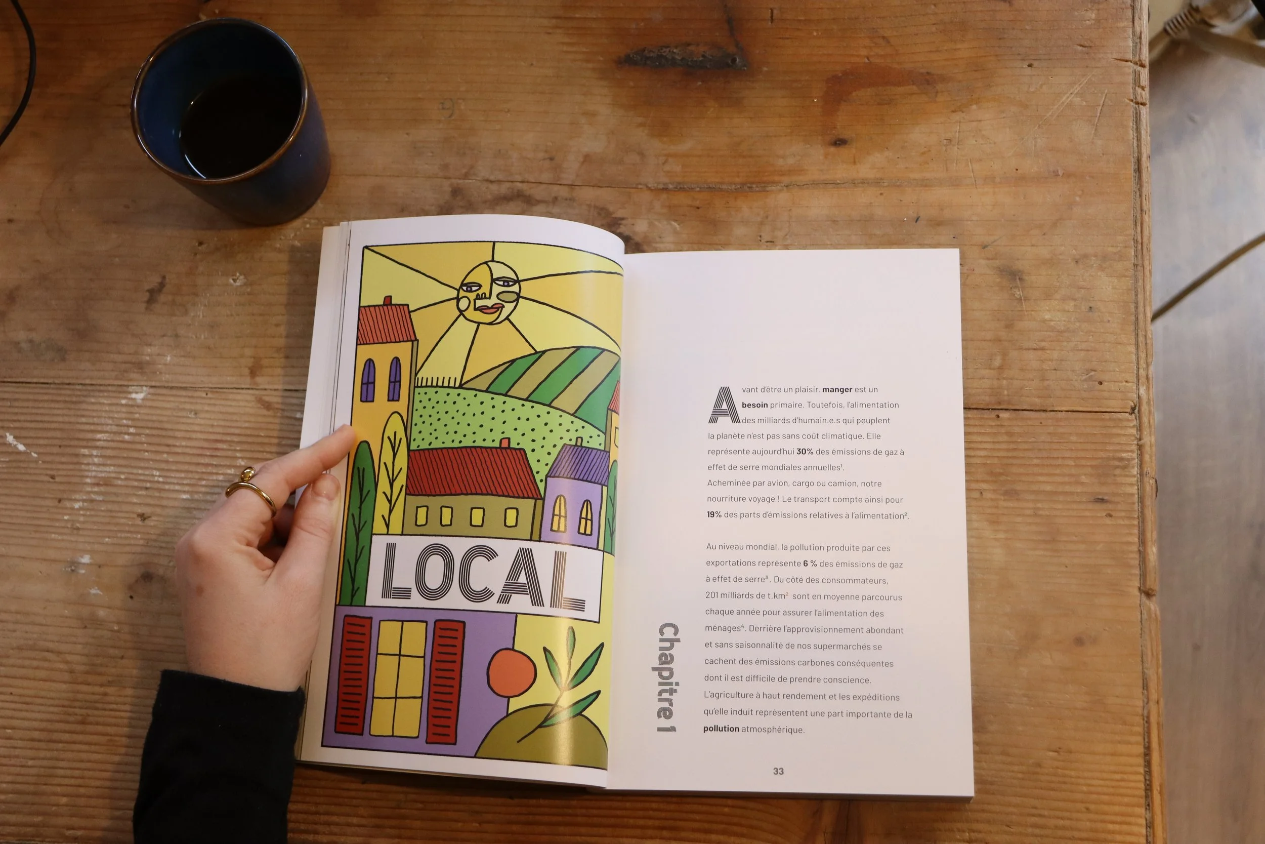 Un livre ouvert affichant une illustration colorée avec des maisons, un soleil souriant, et le mot "LOCAL" écrit en gros. On voit une main tenant le livre, un verre de boisson à gauche, le tout sur une table en bois.