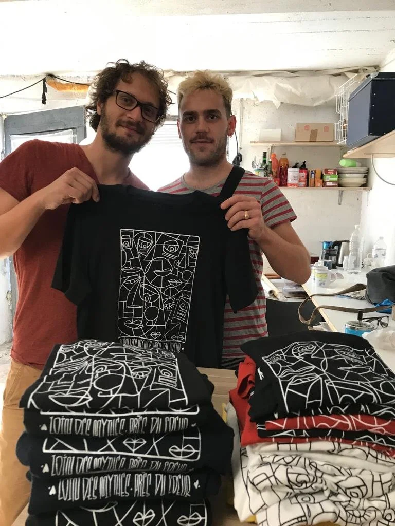 Deux hommes posent dans une cuisine, tenant un t-shirt noir avec un design abstrait blanc. Plusieurs autres t-shirts à motifs similaires sont empilés sur une table devant eux.