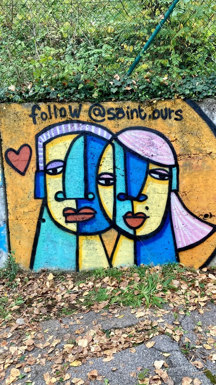 Une fresque artistique représentant deux visages stylisés de femmes, avec des couleurs vives comme le jaune, le bleu et le rose, et un message encourageant à suivre un compte Instagram '@soint.ours'.