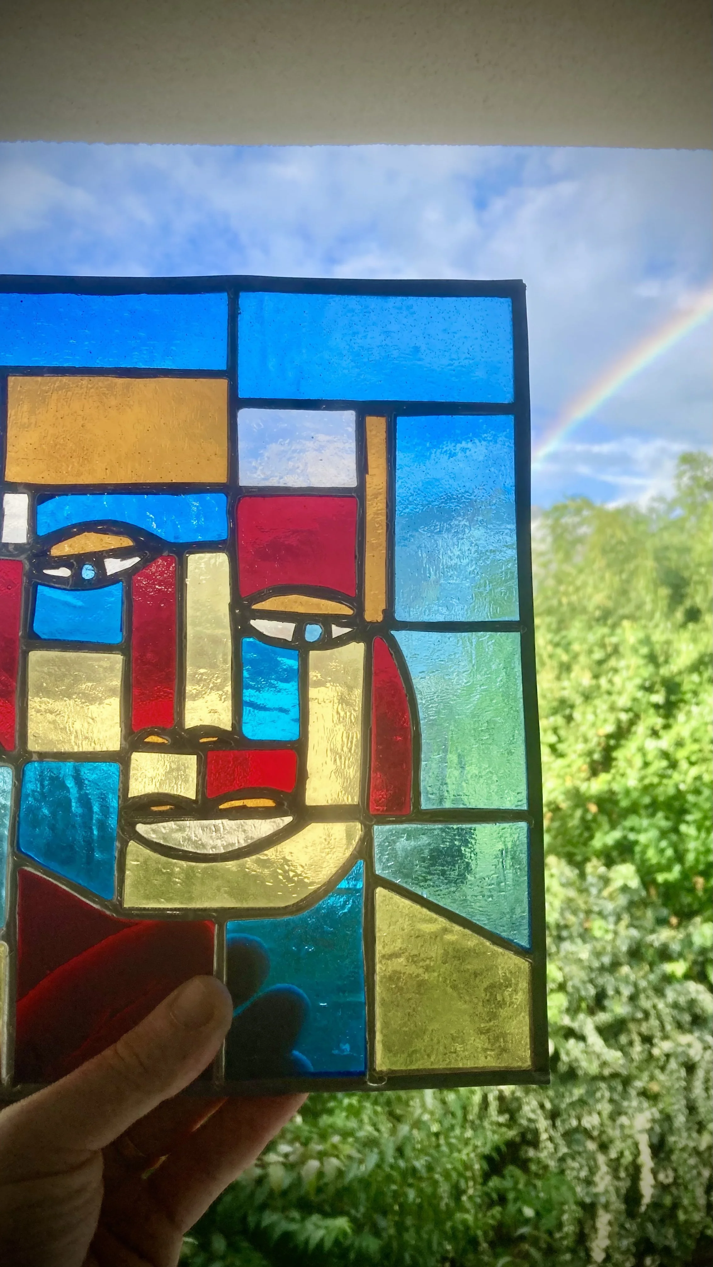 Vitrail représentant un visage avec des couleurs vives telles que rouge, bleu, jaune et vert, avec un ciel bleu et un arc-en-ciel visible en arrière-plan.