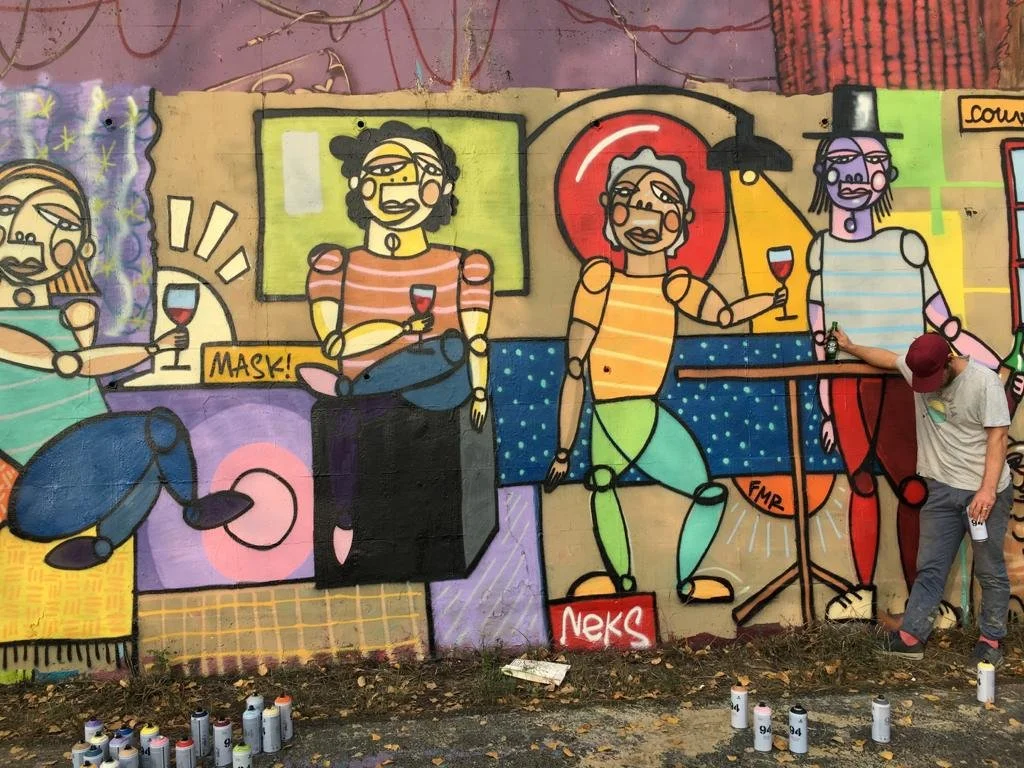 Une fresque murale colorée représentant des personnages stylisés, certains tenant un verre de vin, avec un artiste en train de peindre à droite.