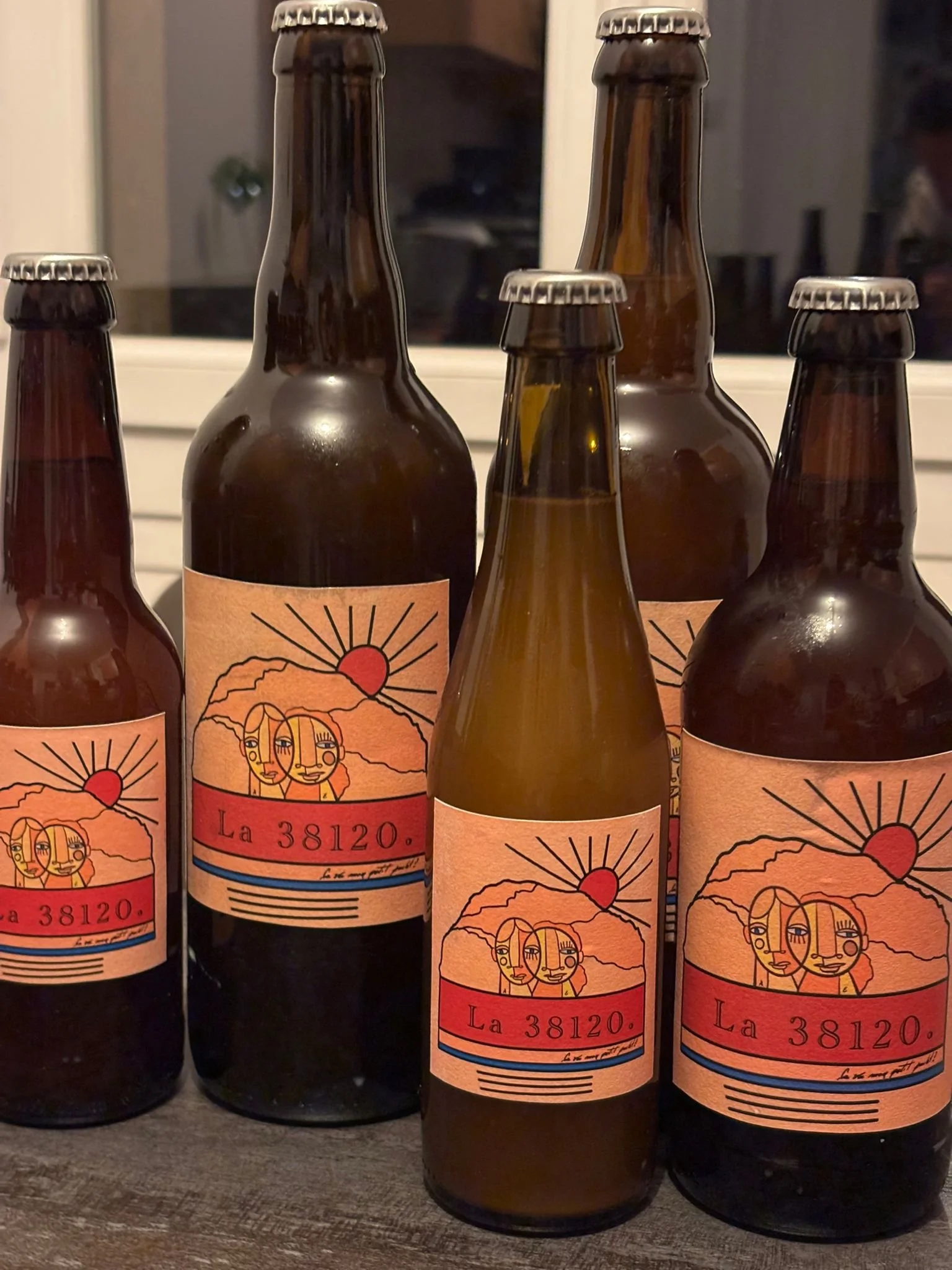 Cinq bouteilles de bière avec une étiquette rose-orange représentant deux visages de femmes, un soleil et une montagne, portant l'inscription 'La 38120'.