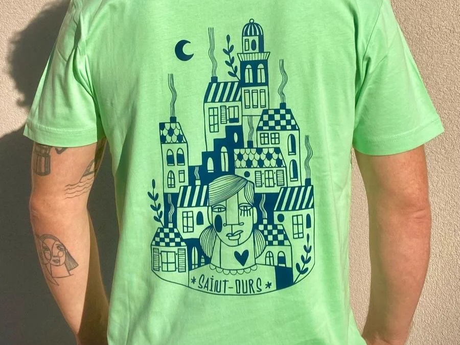 T-shirt vert avec une illustration de maisons stylisées, d'une femme, de feuilles, d'une lune et d'une étoile, avec le texte « Salut Ours » en bas.