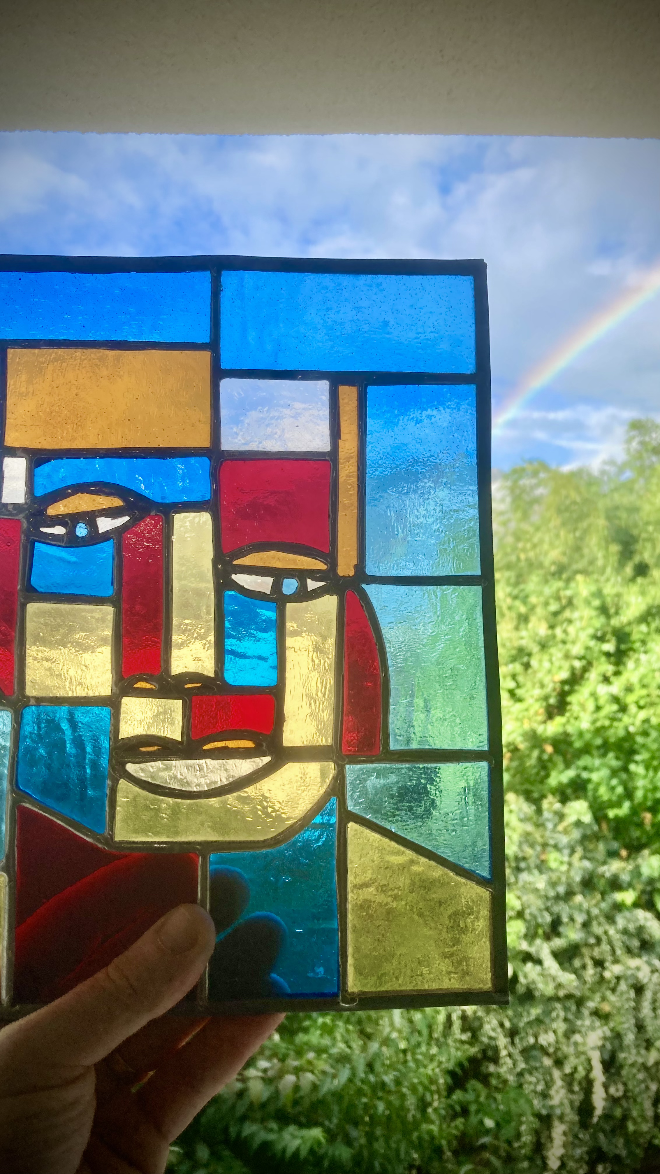 Une vitre teintée représentant un portrait stylisé d'une femme, avec un fond de ciel bleu, nuages et un arc-en-ciel visible à l'extérieur.