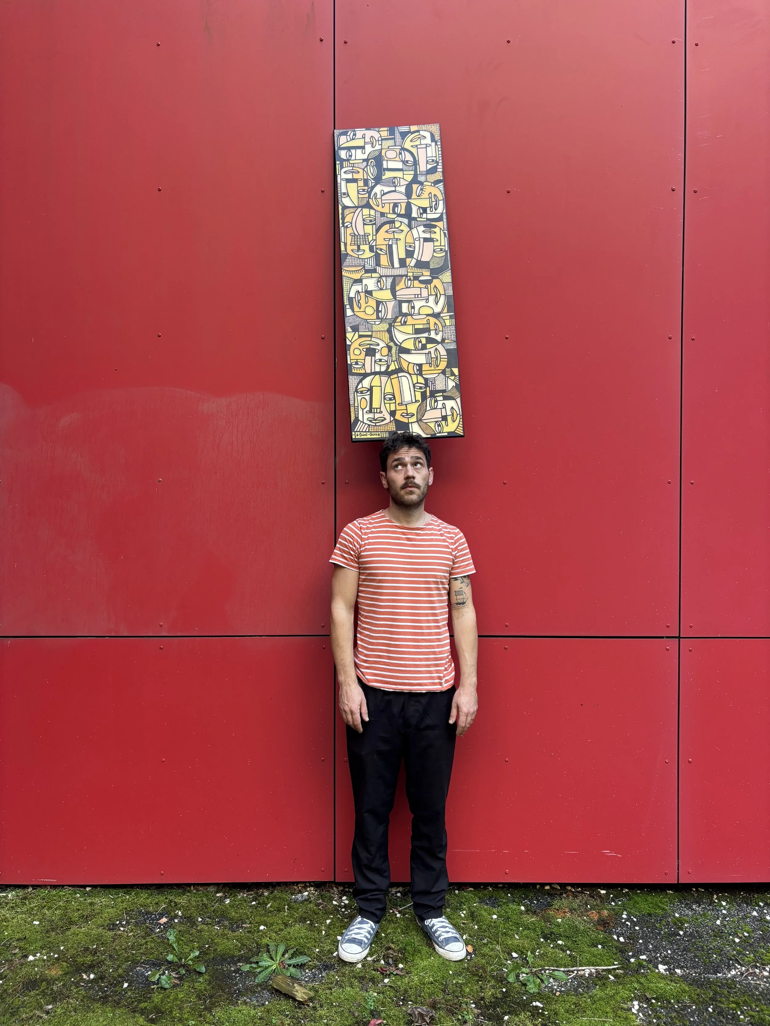 Un homme debout devant un mur rouge, portant un t-shirt à rayures rouges et blanches et un pantalon noir, avec un tableau mural suspendu au-dessus de sa tête.