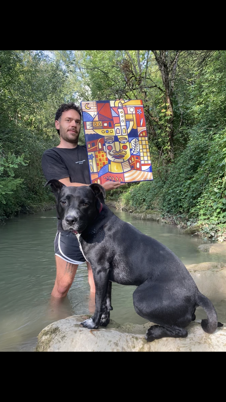 Un homme avec un chien noir dans une rivière entourée de verdure, tenant une peinture colorée avec un visage stylisé et des bâtiments abstraits.