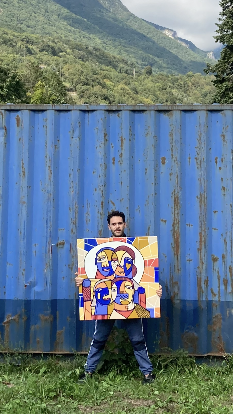 Un homme debout dans un espace extérieur herbeux, tenant une œuvre d'art colorée représentant plusieurs visages stylisés, avec des montagnes verdoyantes en arrière-plan et une grande structure en métal bleue derrière lui.