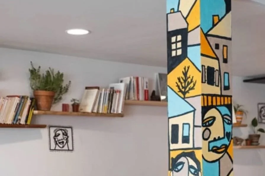 Une pièce intérieure avec des étagères remplies de livres, une plante en pot et une cloison décorative avec une peinture abstraite colorée.