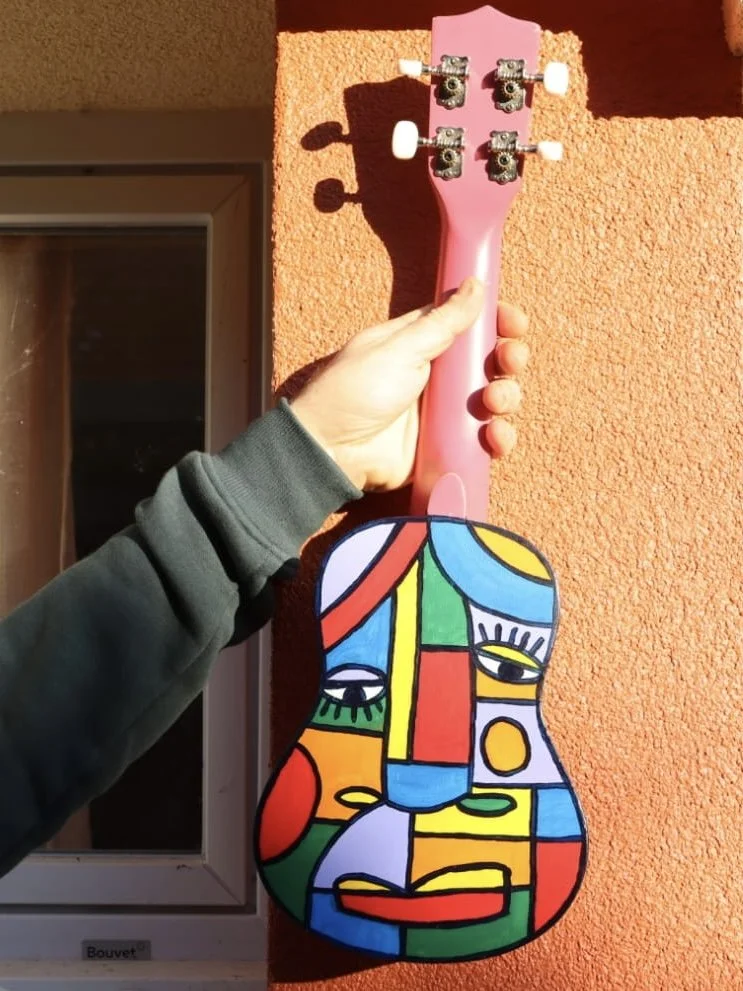 Une main tient une tête de guitare peinte avec un motif abstrait coloré de style cubiste, posée contre un mur en brique orange, avec un manche de guitare rose au-dessus.