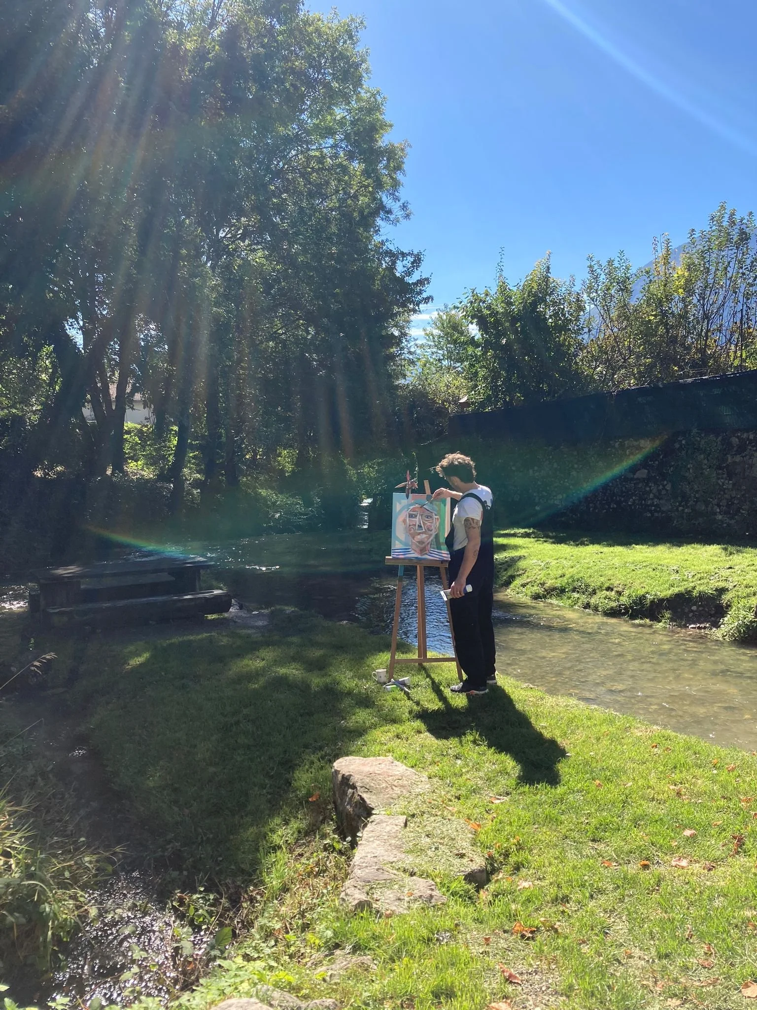 Un artiste peint un portrait facial sur une toile à chevalet dans un parc en plein soleil, avec des arbres, une rivière, et une grande roche devant lui.