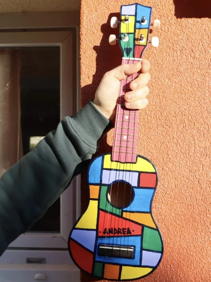 Une guitare en bois décorée avec un motif coloré ressemblant à une œuvre de Mondrian, portée par une personne en sweatshirt gris, avec un nom "ANDREA" écrit sur le corps de la guitare, devant un mur orange.