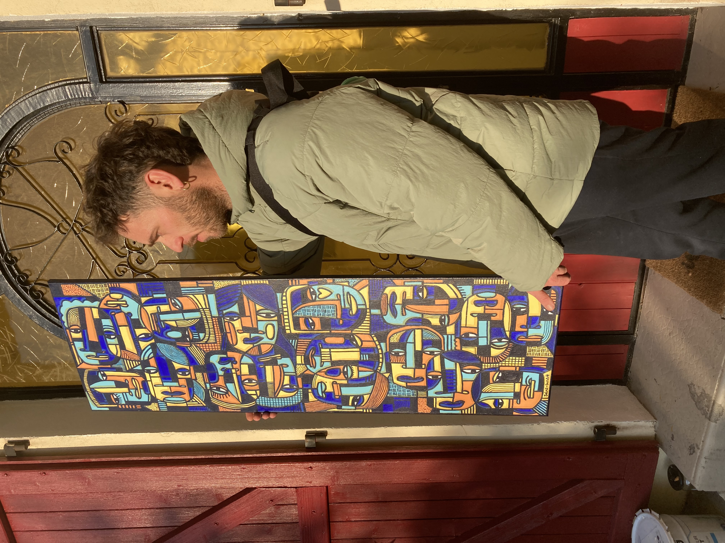 Un homme tenant une peinture colorée abstracte, dans un décor intérieur avec portes en bois rouge et décorations en métal.