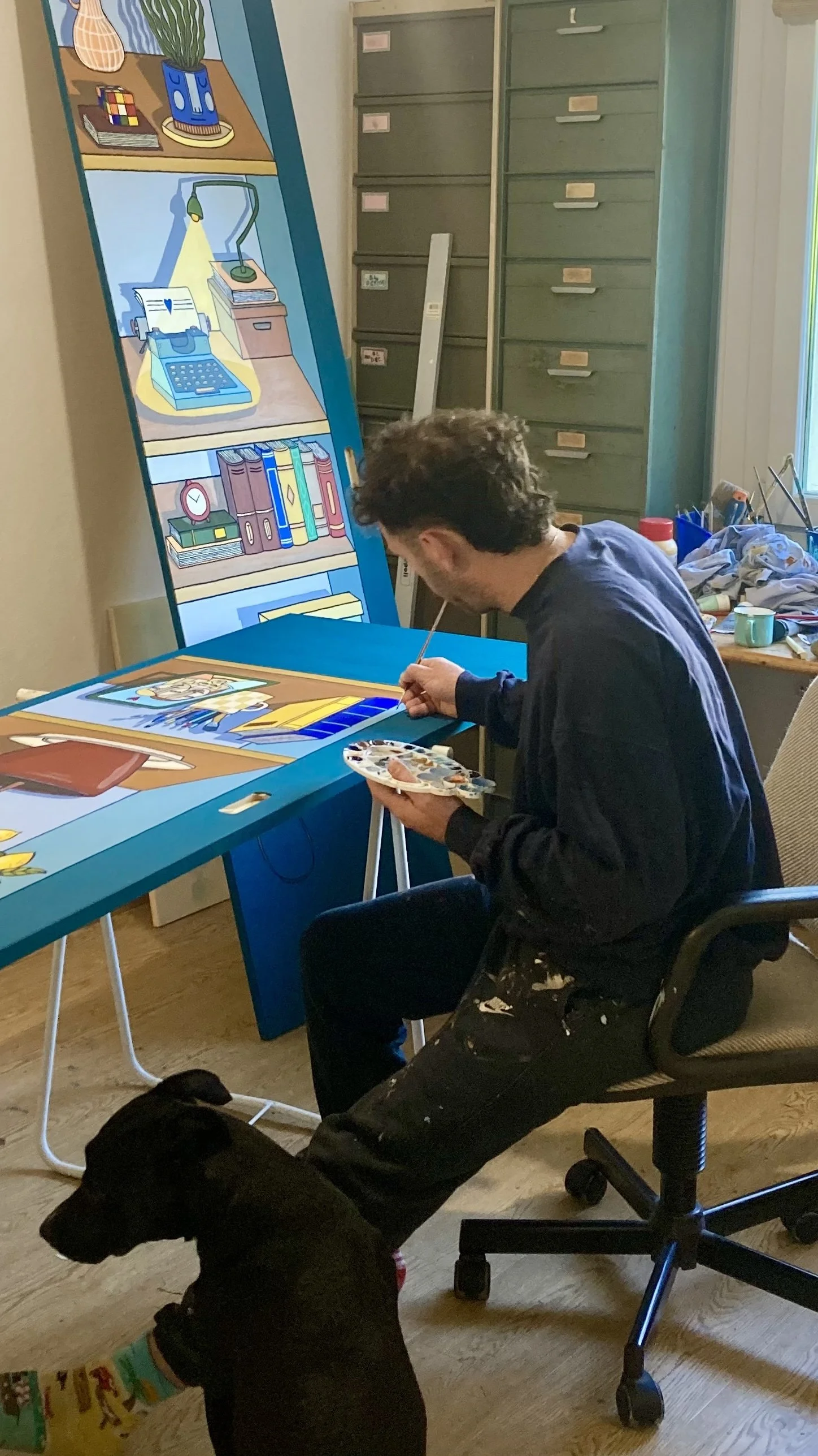 Une personne en train de peindre une illustration pour une peinture murale ou un panneau en bois. Il y a un chien noir assis à côté d'elle.