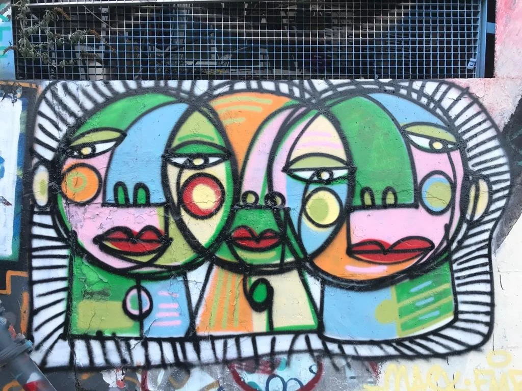 Graffiti coloré représentant plusieurs visages stylisés avec des traits abstraits, entourés d'une bordure noire ondulée.