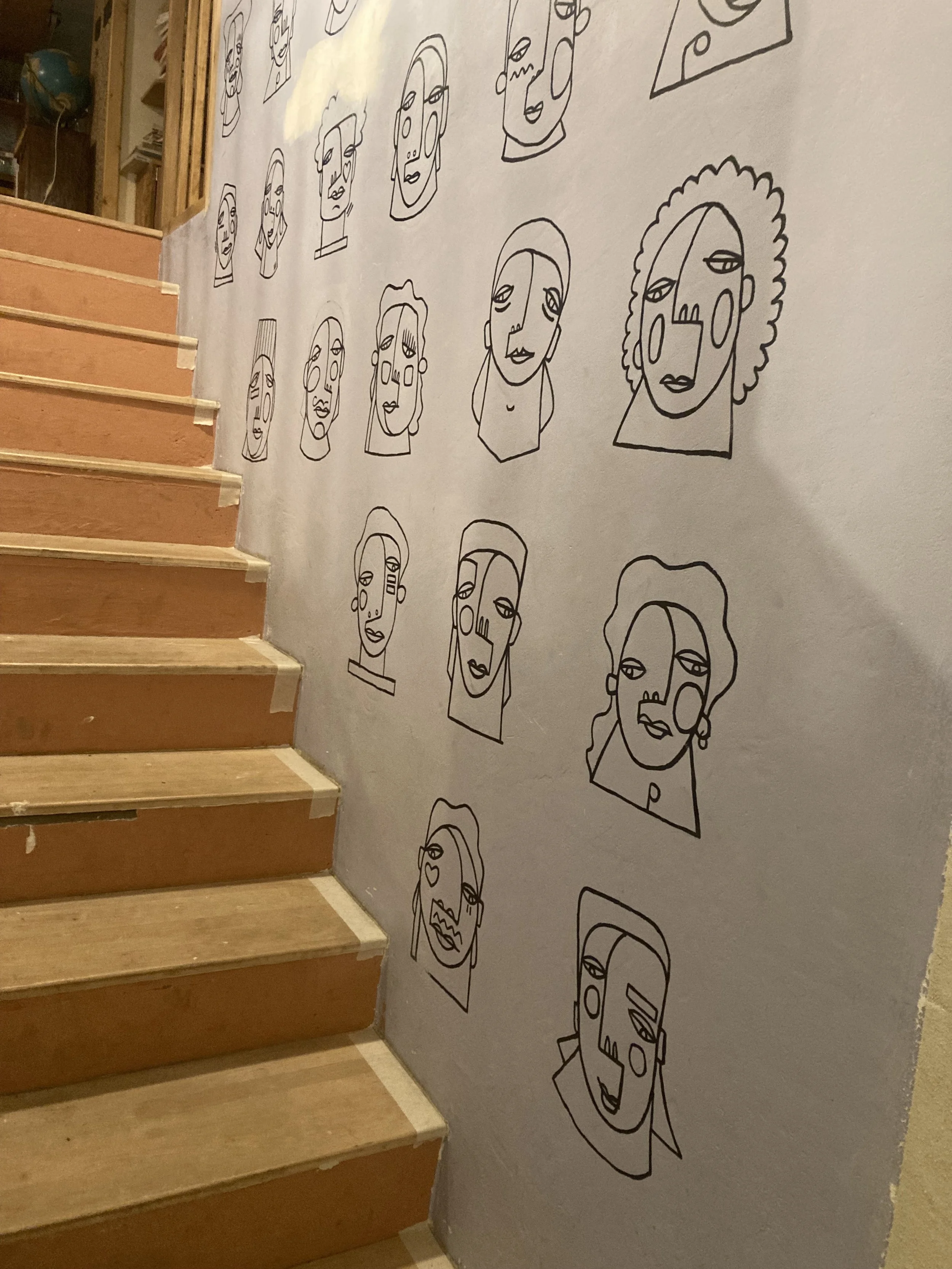 Dix-huit dessins de têtes de femmes faites à la ligne, décorant un mur à côté d'un escalier en bois.