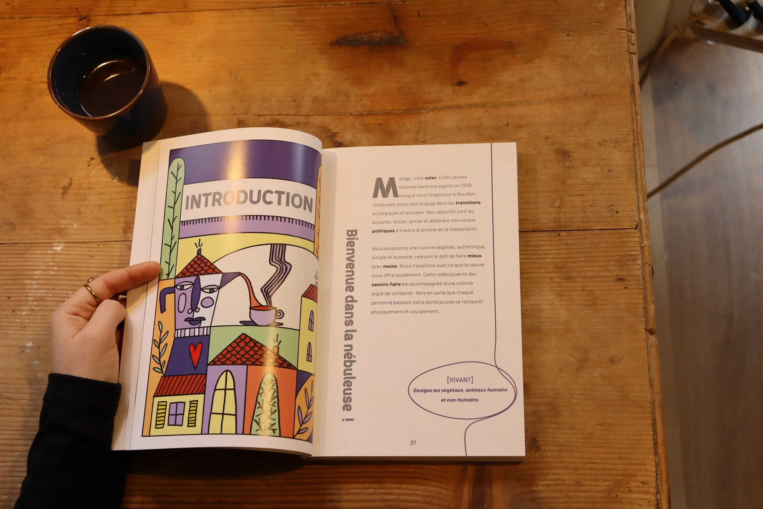 Un livre ouvert sur une table en bois, montrant une page d'introduction avec une illustration colorée d'une maison, arbre et personnage stylisé, et un texte en français. Une main tient la page à gauche, et une tasse vide est placée à côté du livre.