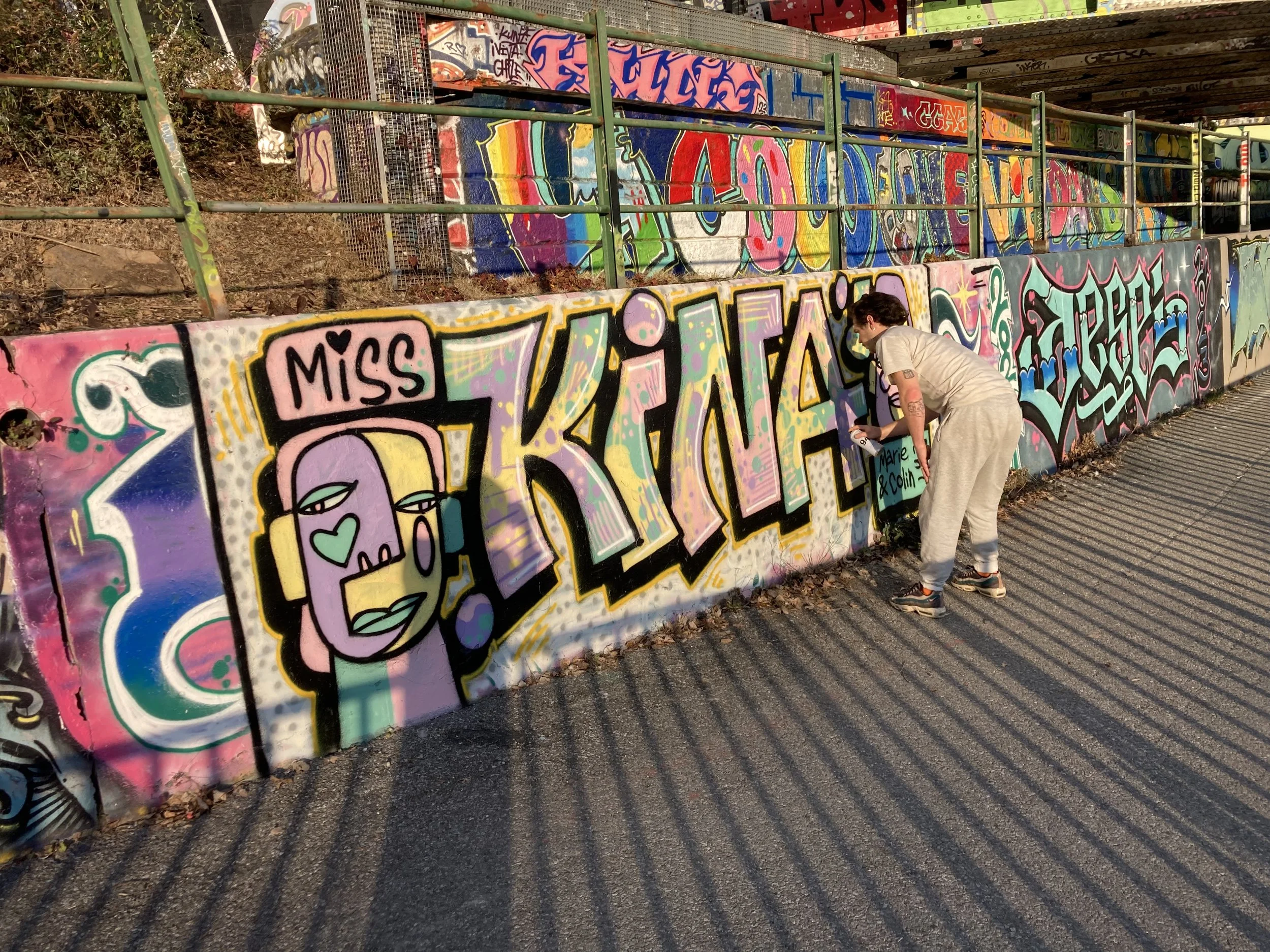 Une personne en train de peindre un graffiti coloré sur un mur en béton. Le graffiti comprend le mot 'Miss' et un visage stylisé avec des couleurs pastel.