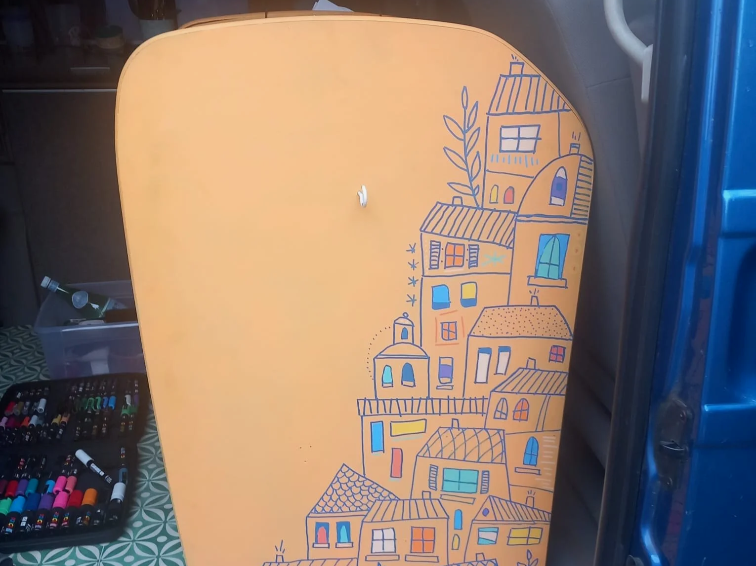 Table jaune décorée avec un dessin de maisons colorées et de végétation, entourée de stylos et de matériel d'art.