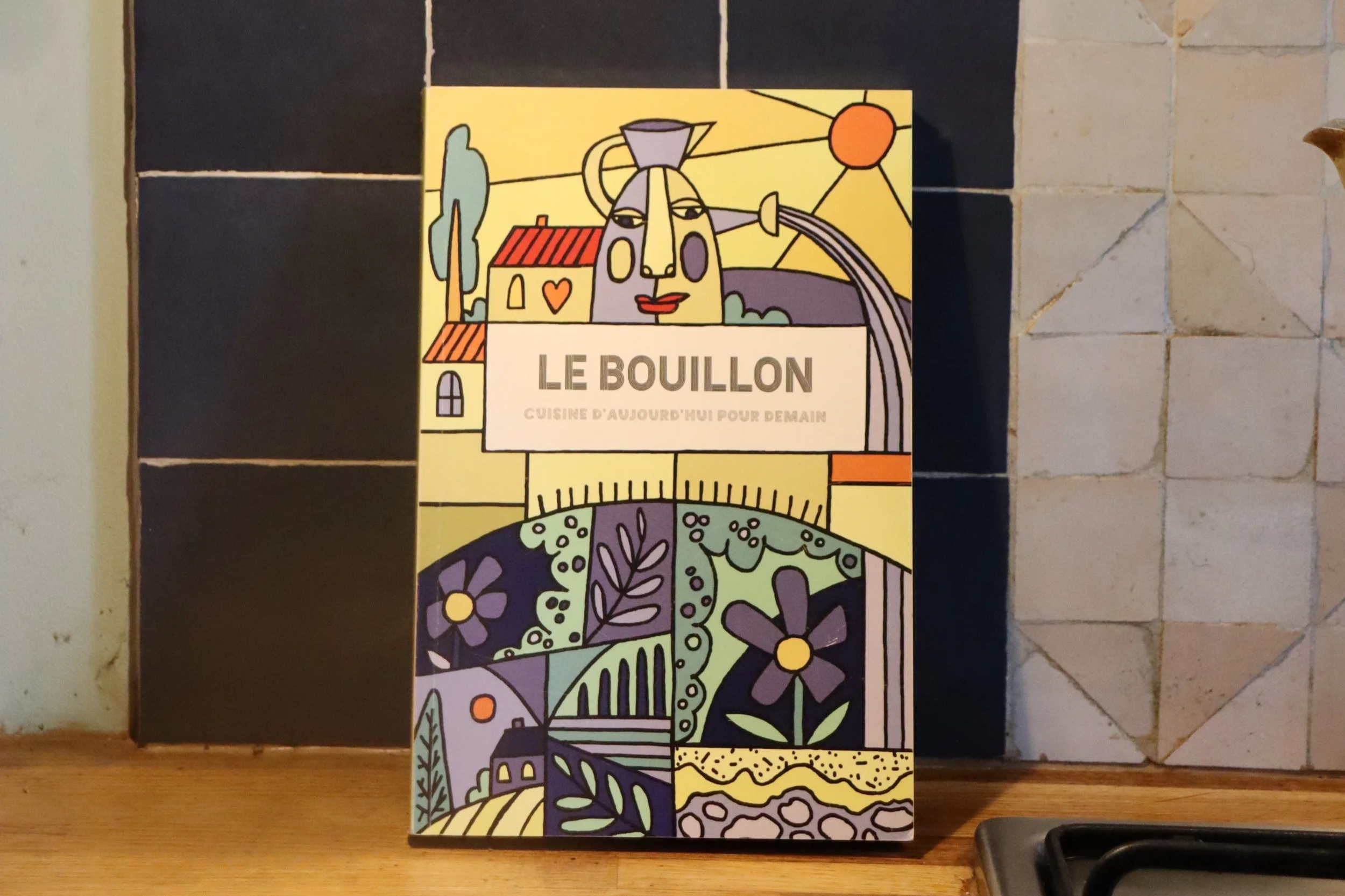 Une affiche colorée avec un dessin de style abstrait représentant une maison, un arbre, des fleurs et un ciel avec le soleil. Le texte sur l'affiche indique 'Le Bouillon' et 'Cuisine d'aujourd'hui pour demain'.