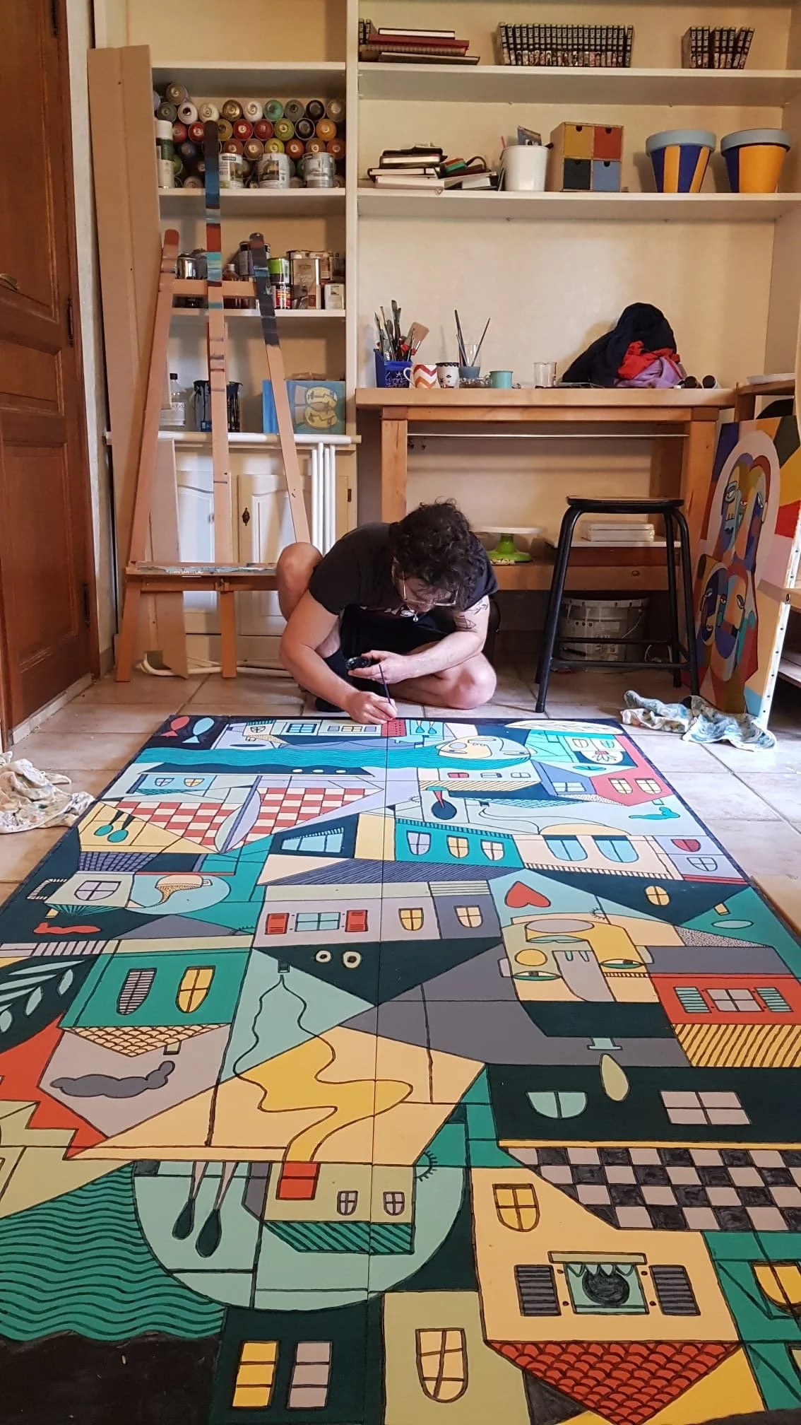Un homme peint un puzzle géant représentant un village avec des maisons colorées et des éléments de paysage, dans un atelier avec des étagères et des outils en arrière-plan.